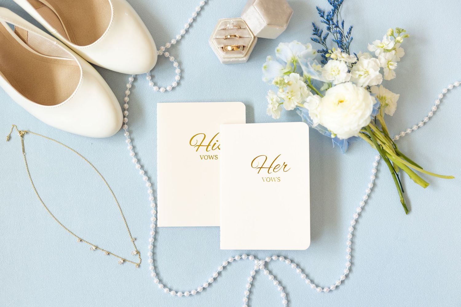 Flat lay of two Her vows books, white bridal shoes, pearl necklace, ring box, and blue floral bouquet on blue background.