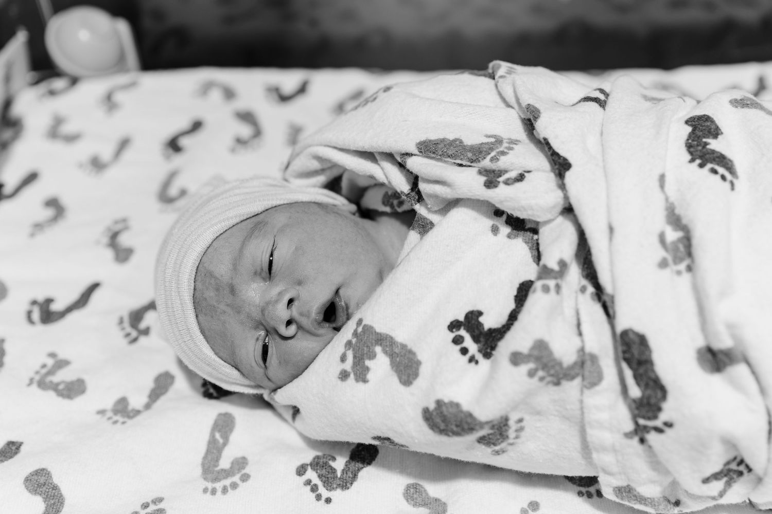 Newborn baby swaddled in a soft blanket with footprint patterns, wearing a knit cap and sleeping peacefully in black and white.