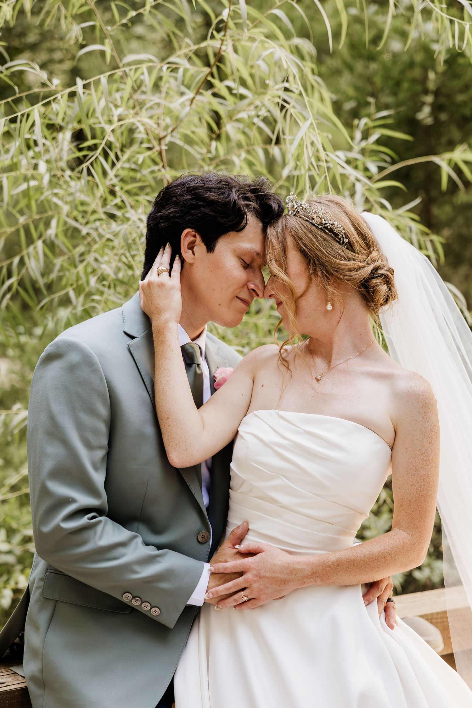 A bride and groom share a romantic moment, touching foreheads, surrounded by lush greenery on their wedding day.