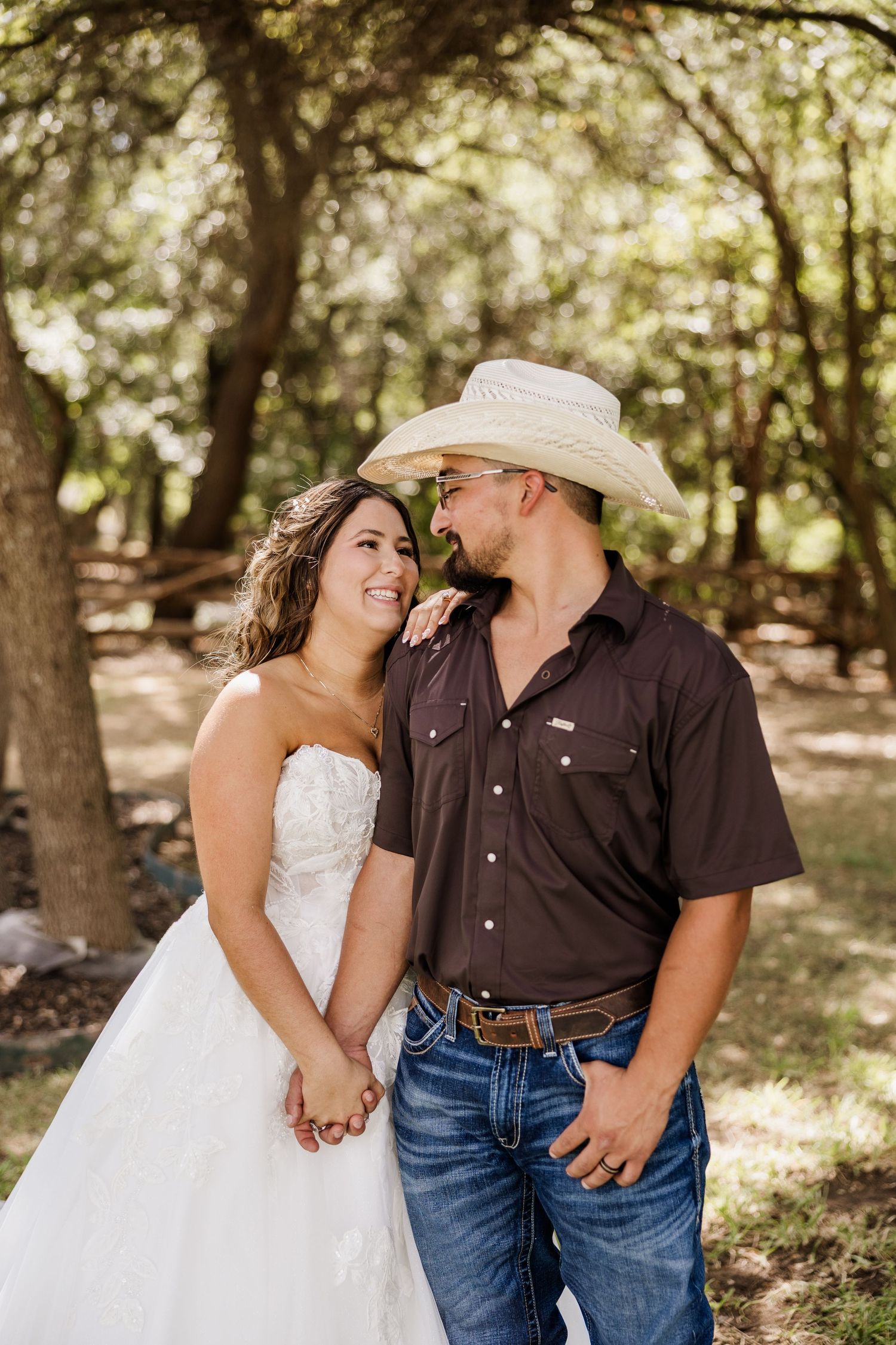 Bride in white strapless gown and groom in brown shirt, jeans, and cowboy hat share a romantic moment outdoors.