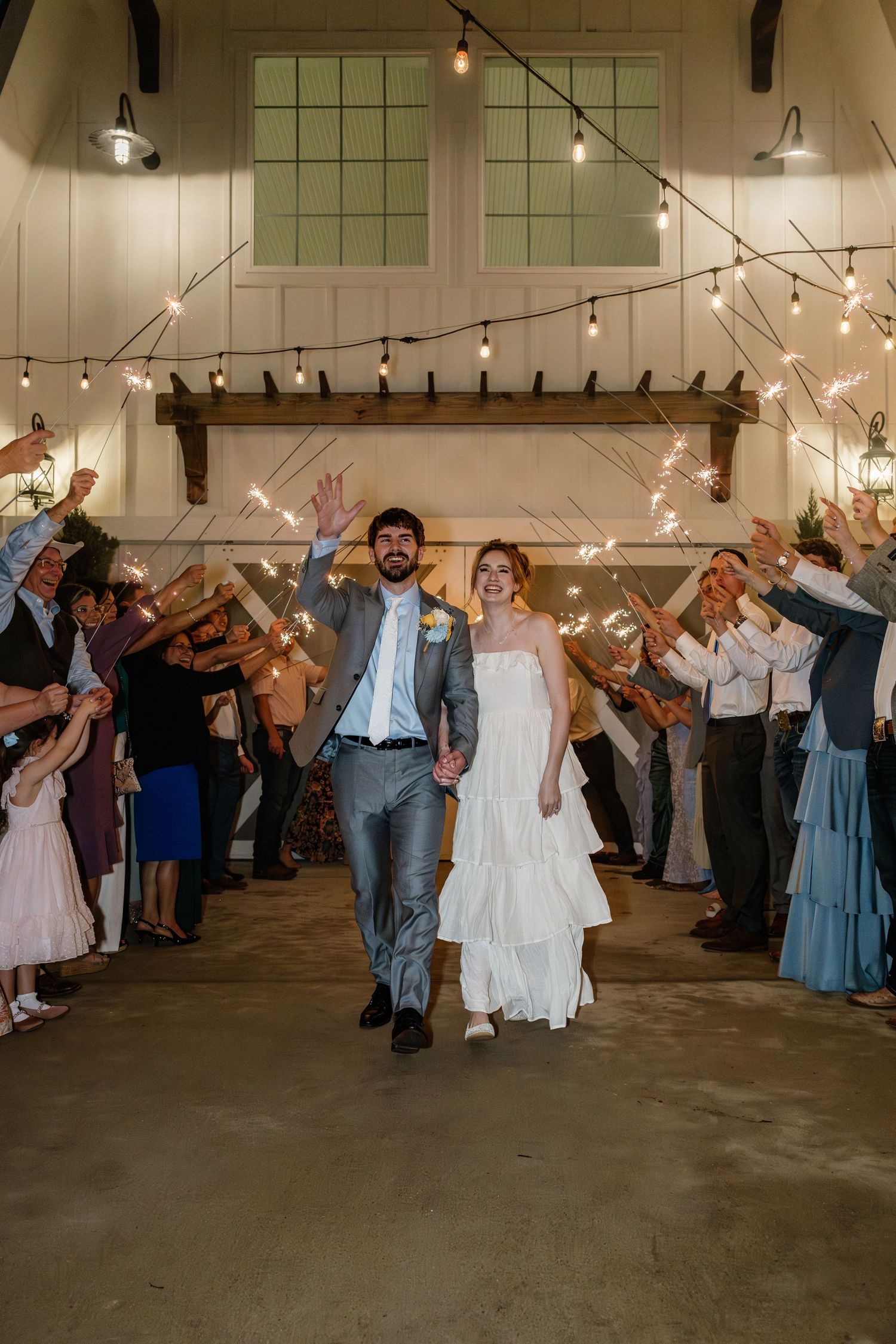 Bride and groom exit wedding venue through sparkler tunnel while guests celebrate in rustic barn setting.