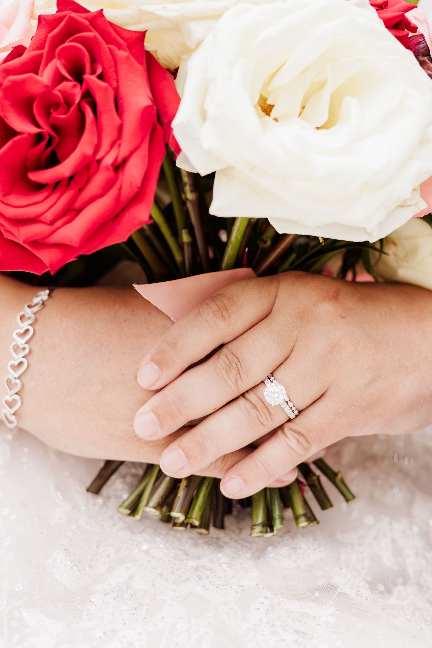 Bride's hands resting on red and white rose bouquet, showcasing diamond engagement ring and delicate silver bracelet.
