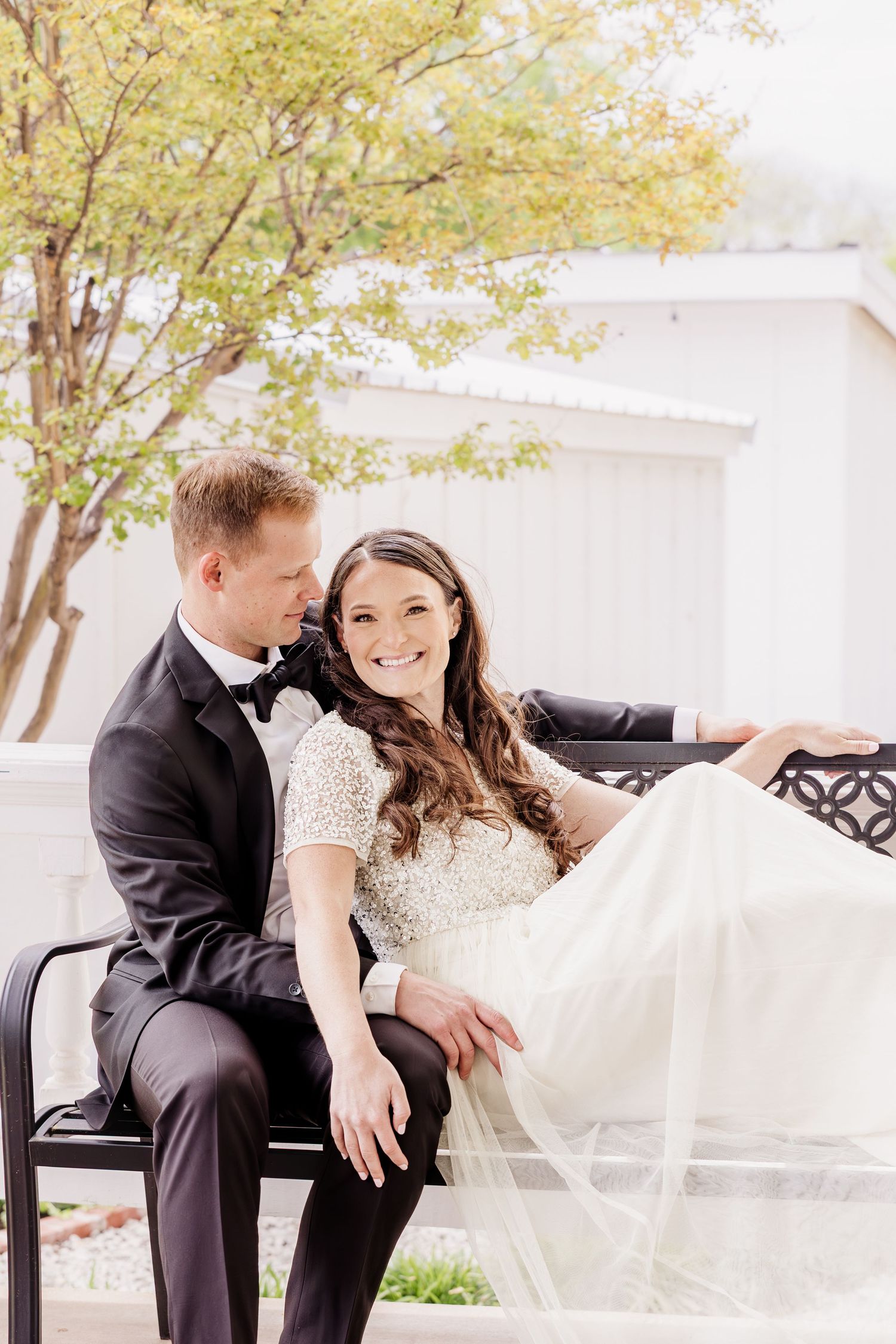 A bride and groom sit together on a bench outdoors, smiling, with the bride in a beaded gown and groom in a black tuxedo.