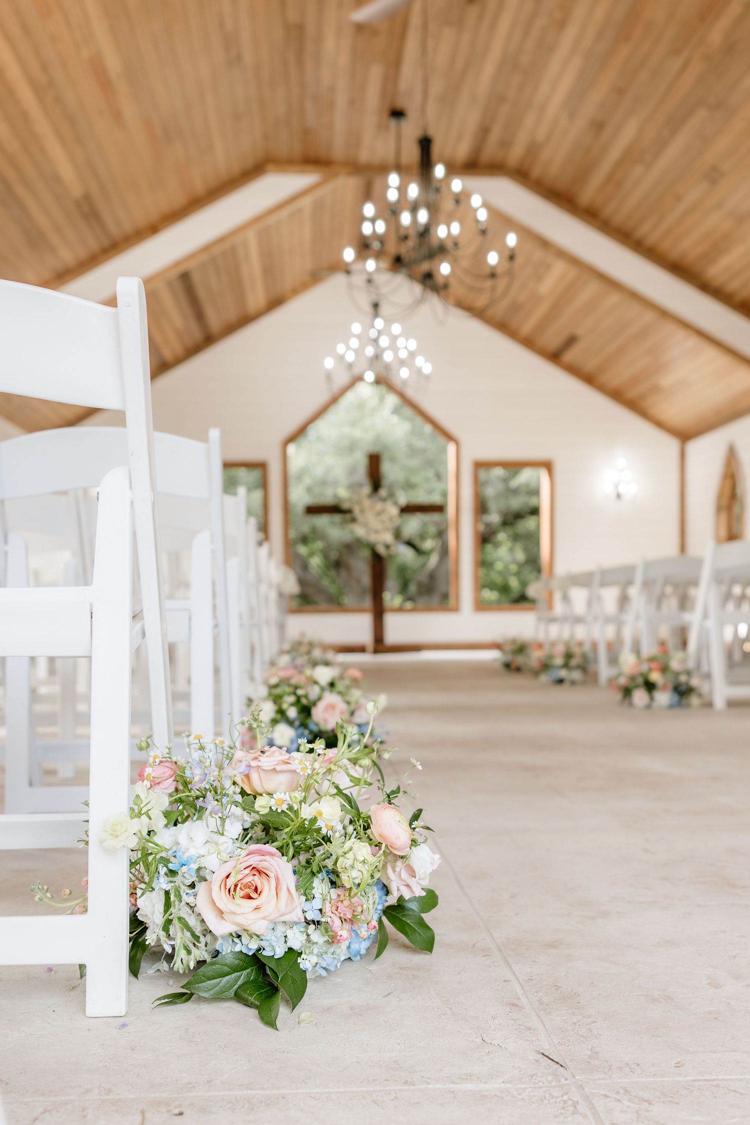 Elegant wedding chapel with white chairs, floral arrangements, wood ceiling, chandelier, and a cross at the altar.
