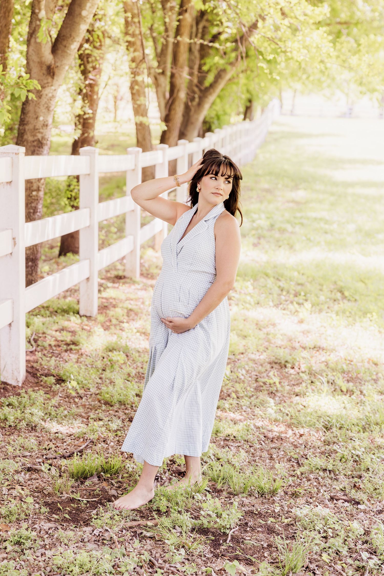 Pregnant woman in light blue dress standing barefoot by white fence among green trees, cradling baby bump outdoors.