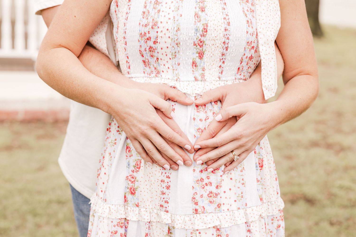 Couple forming heart shape with hands on pregnant belly in floral dress during maternity photo session outdoors.