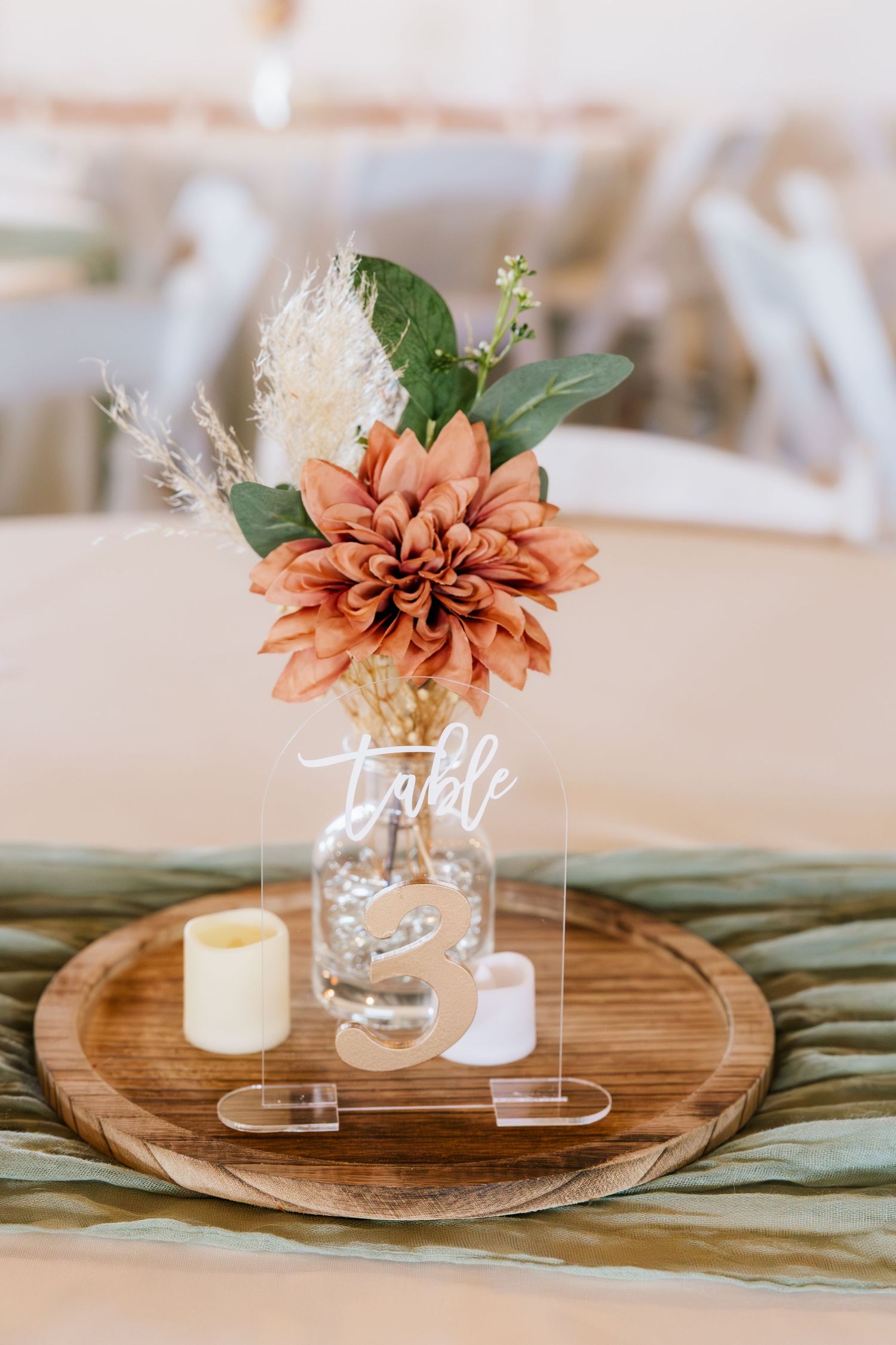 Rustic wedding table centerpiece with terracotta dahlia, dried florals, candles, and acrylic table number on wooden tray.
