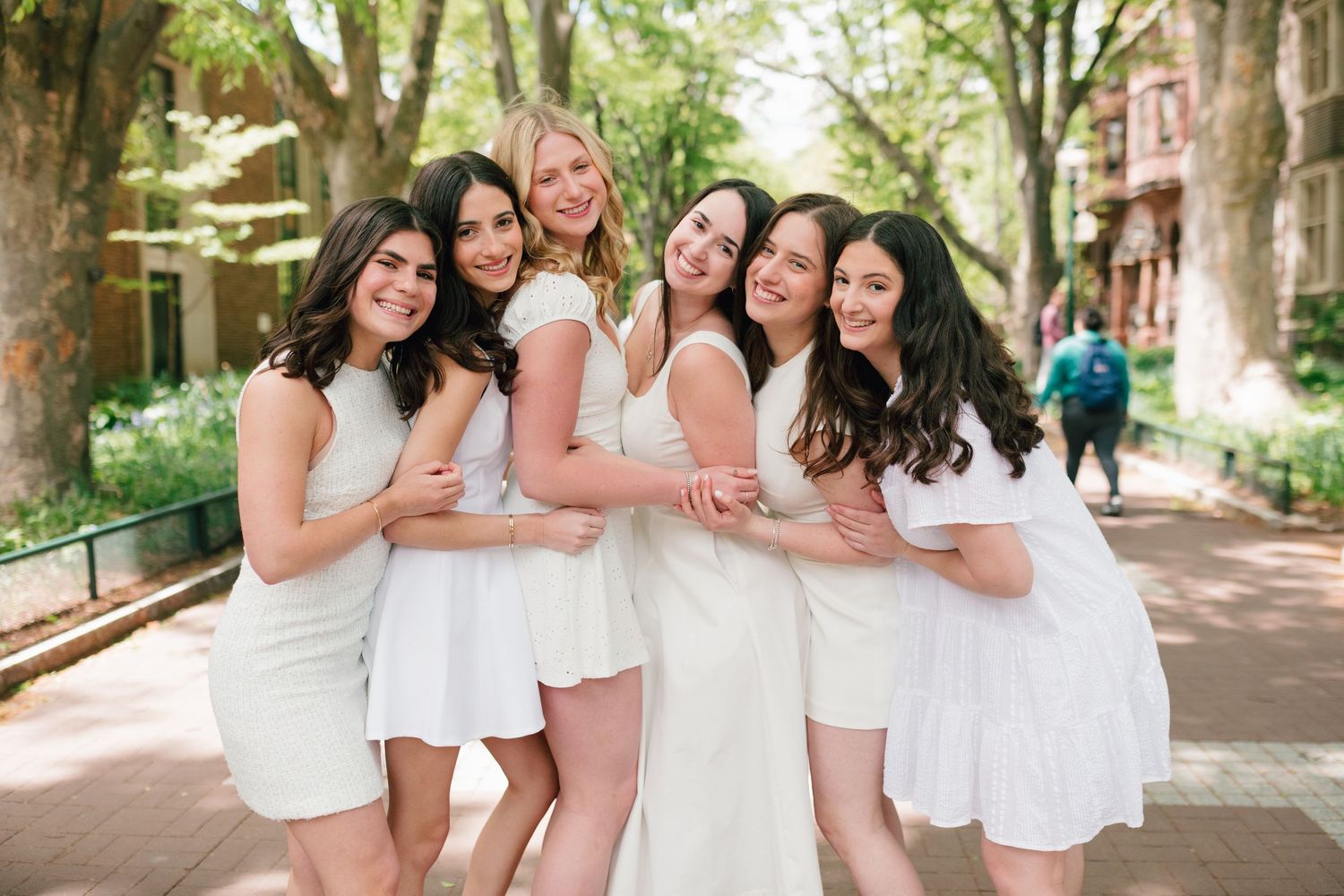 A group in white dresses poses together on a tree-lined path during a celebratory photoshoot.
