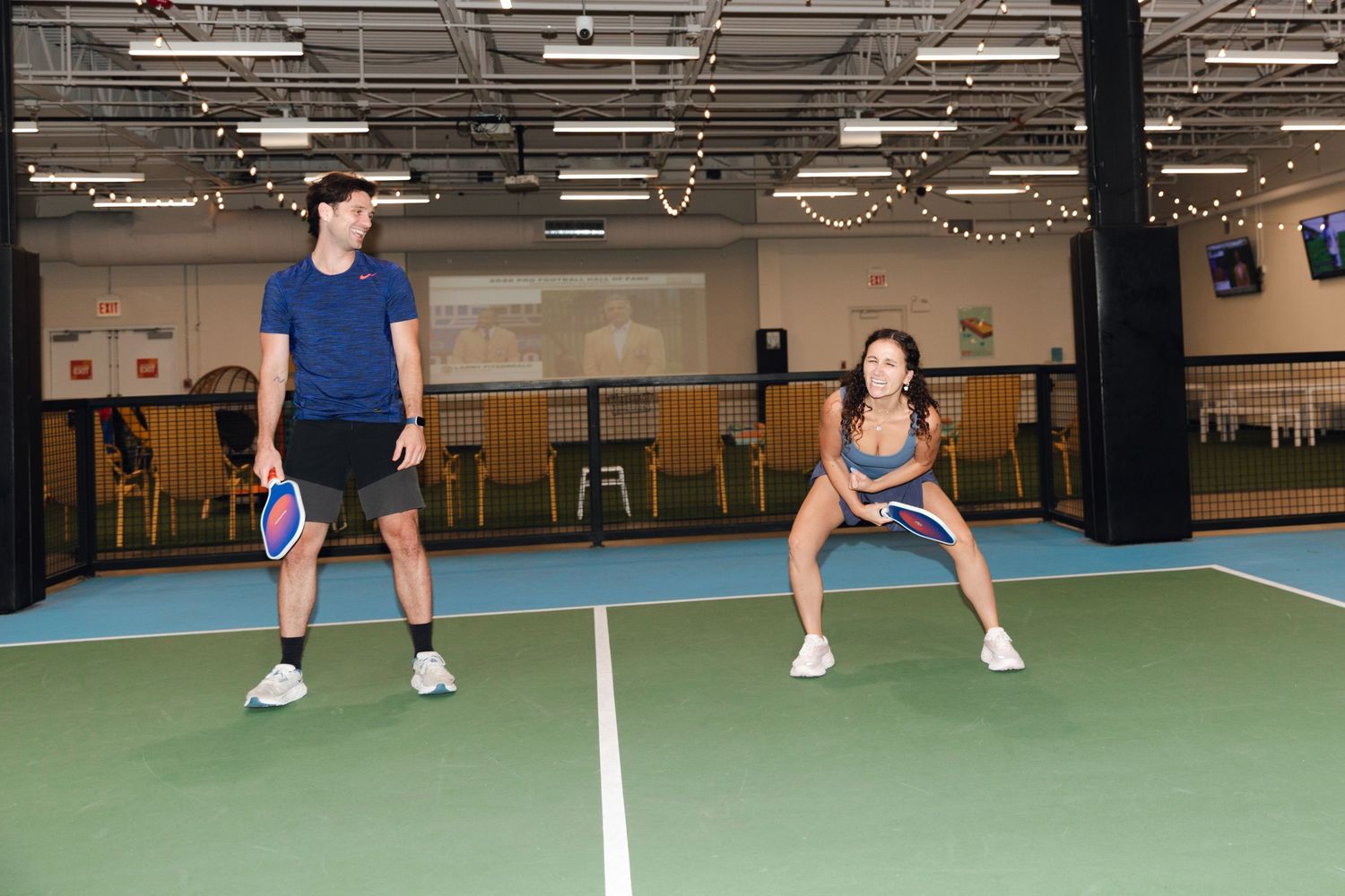 Two players practicing pickleball on an indoor court, holding paddles in athletic stances under bright lights.