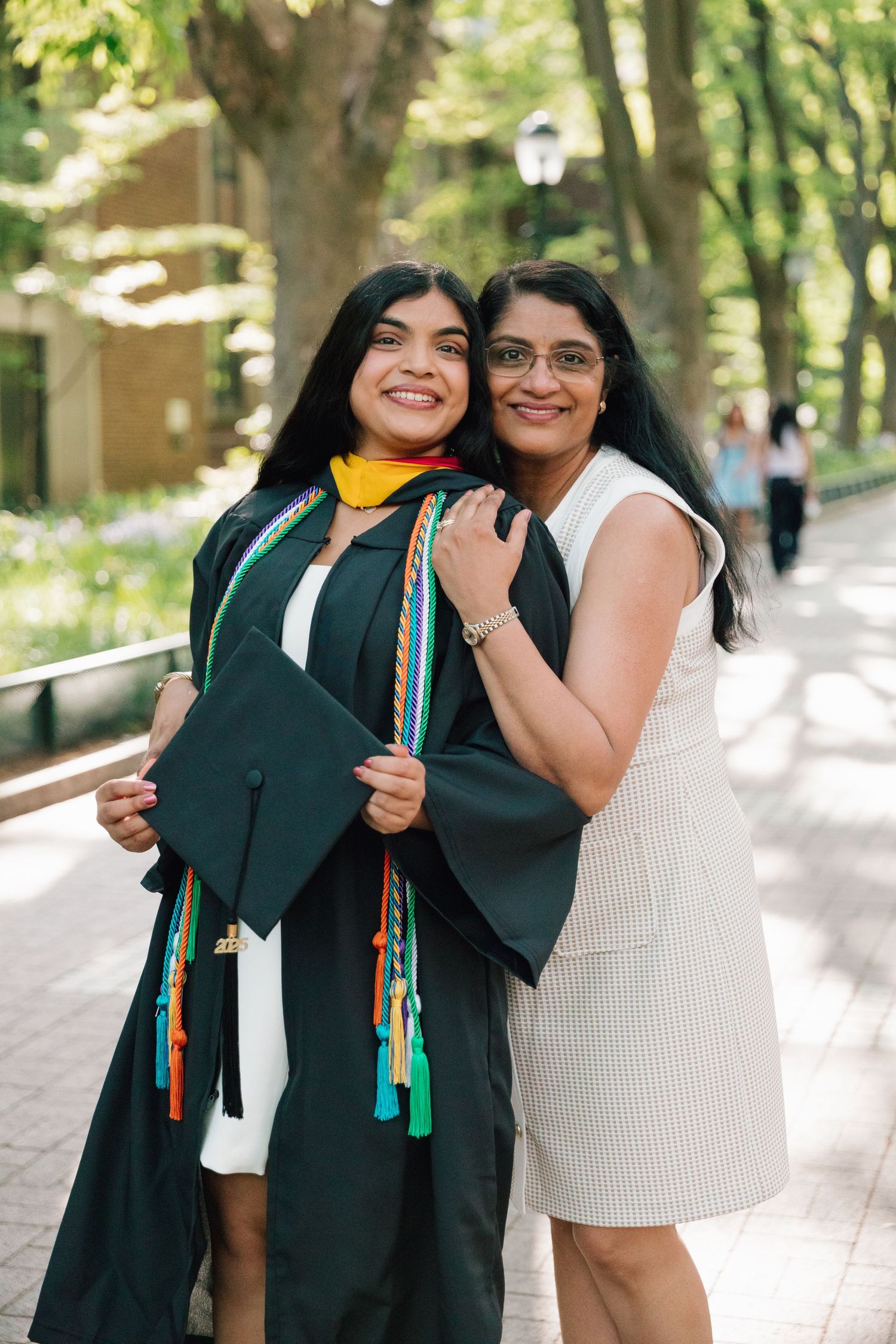 Two people embracing in graduation attire on sunny campus walkway.