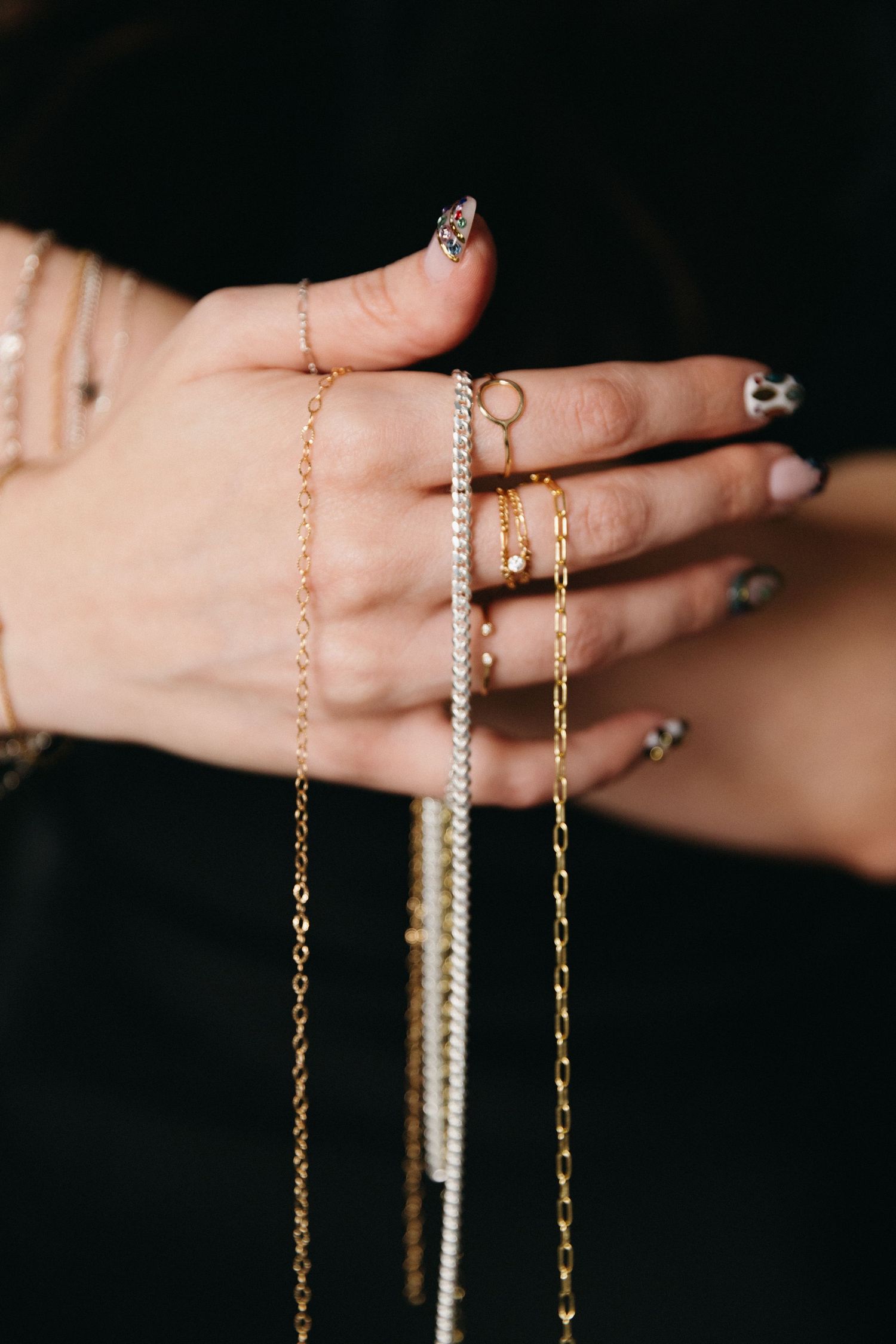 A hand with patterned nails holds multiple delicate gold and silver chain necklaces and wears dainty rings.