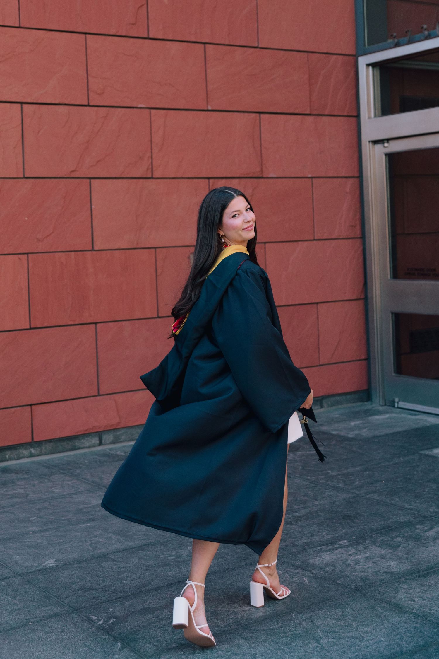 Graduate in black cap and gown poses dramatically against red brick wall.