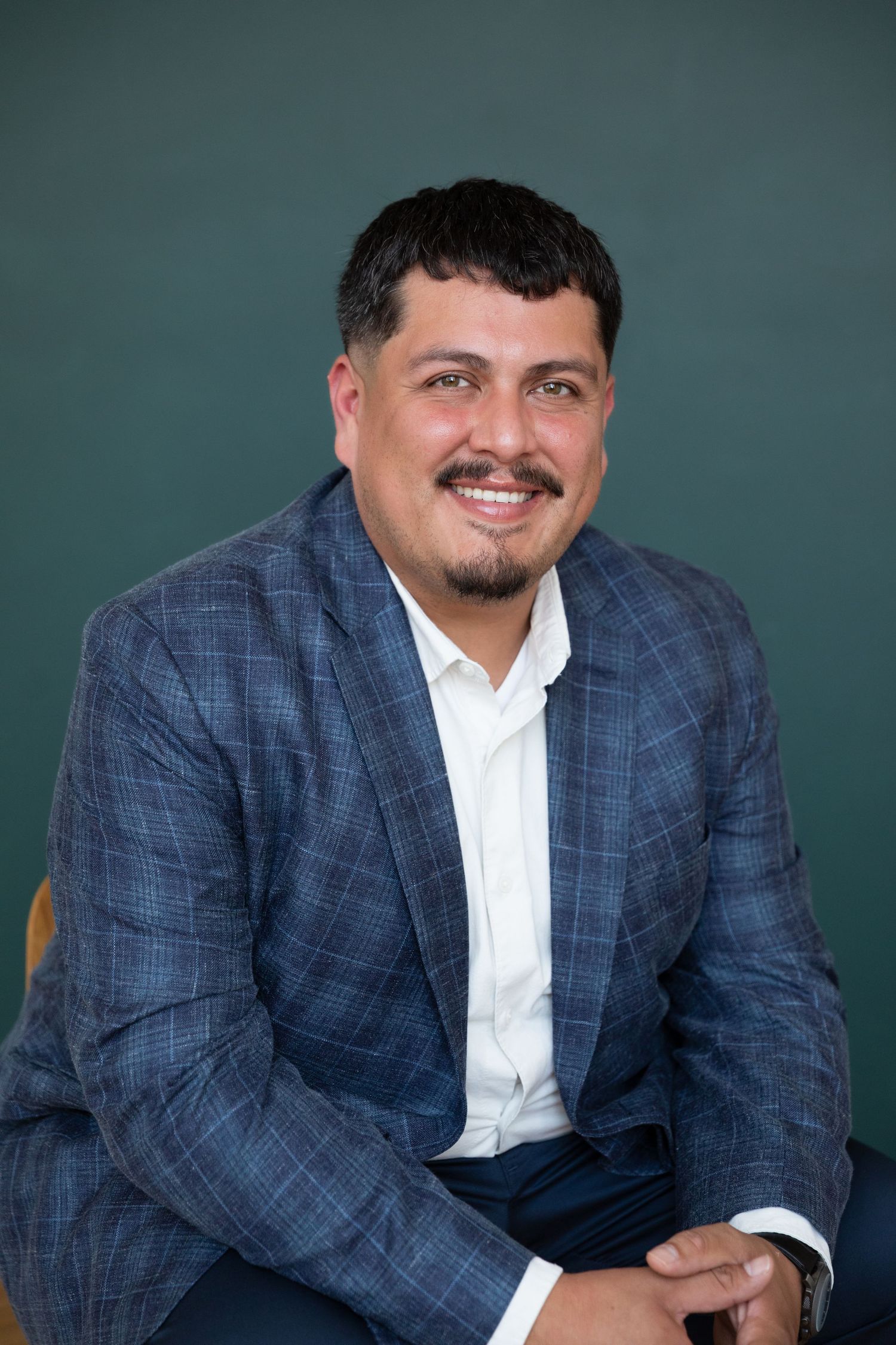 Smiling man in a blue plaid blazer and white shirt seated against a dark teal background.