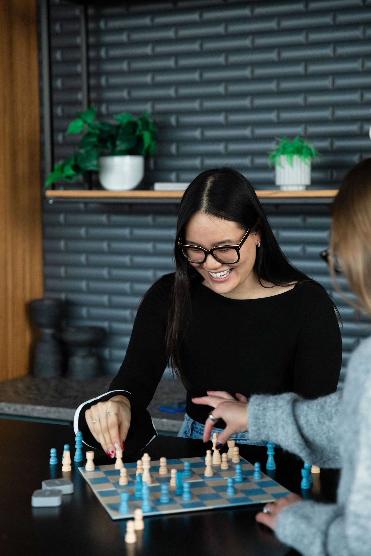 Two women smiling and playing chess on a dark wooden table in a modern cafe with brick wall backdrop.