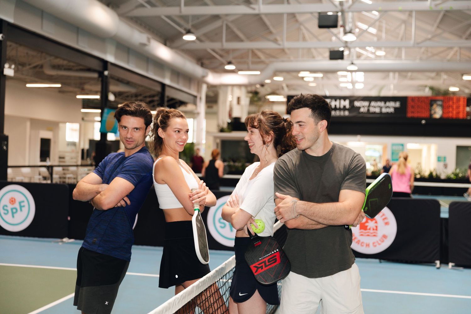 Four players pose at a tennis net inside a modern indoor court, holding rackets with arms crossed.