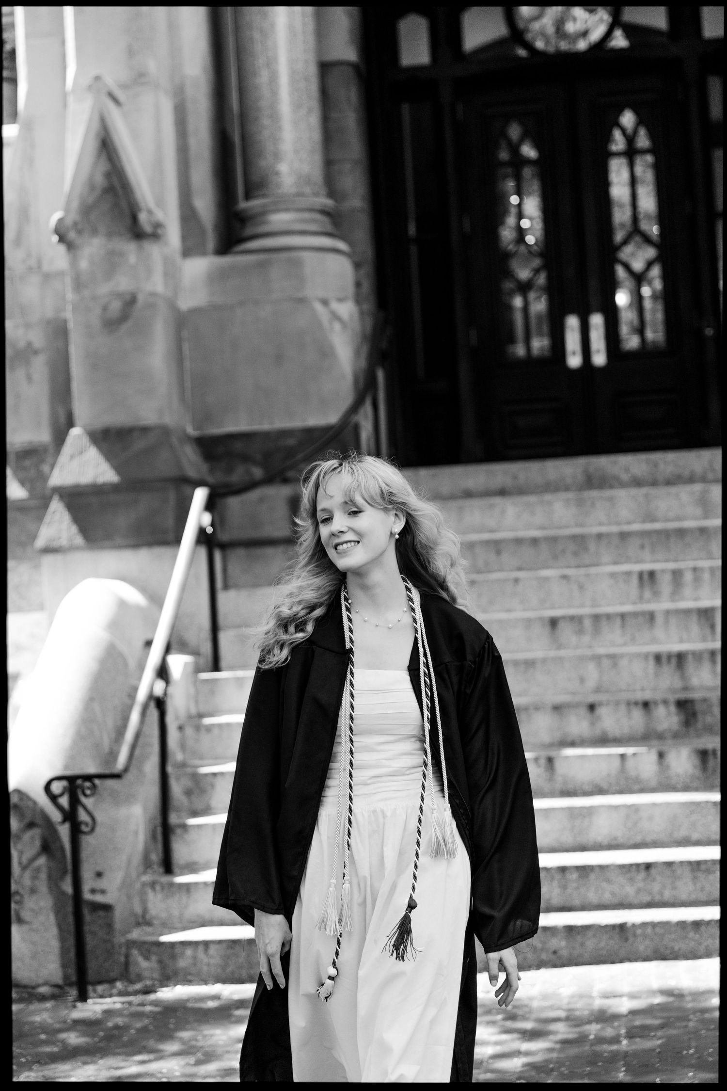 Graduate student in black academic regalia smiling on stone steps of historic university building.