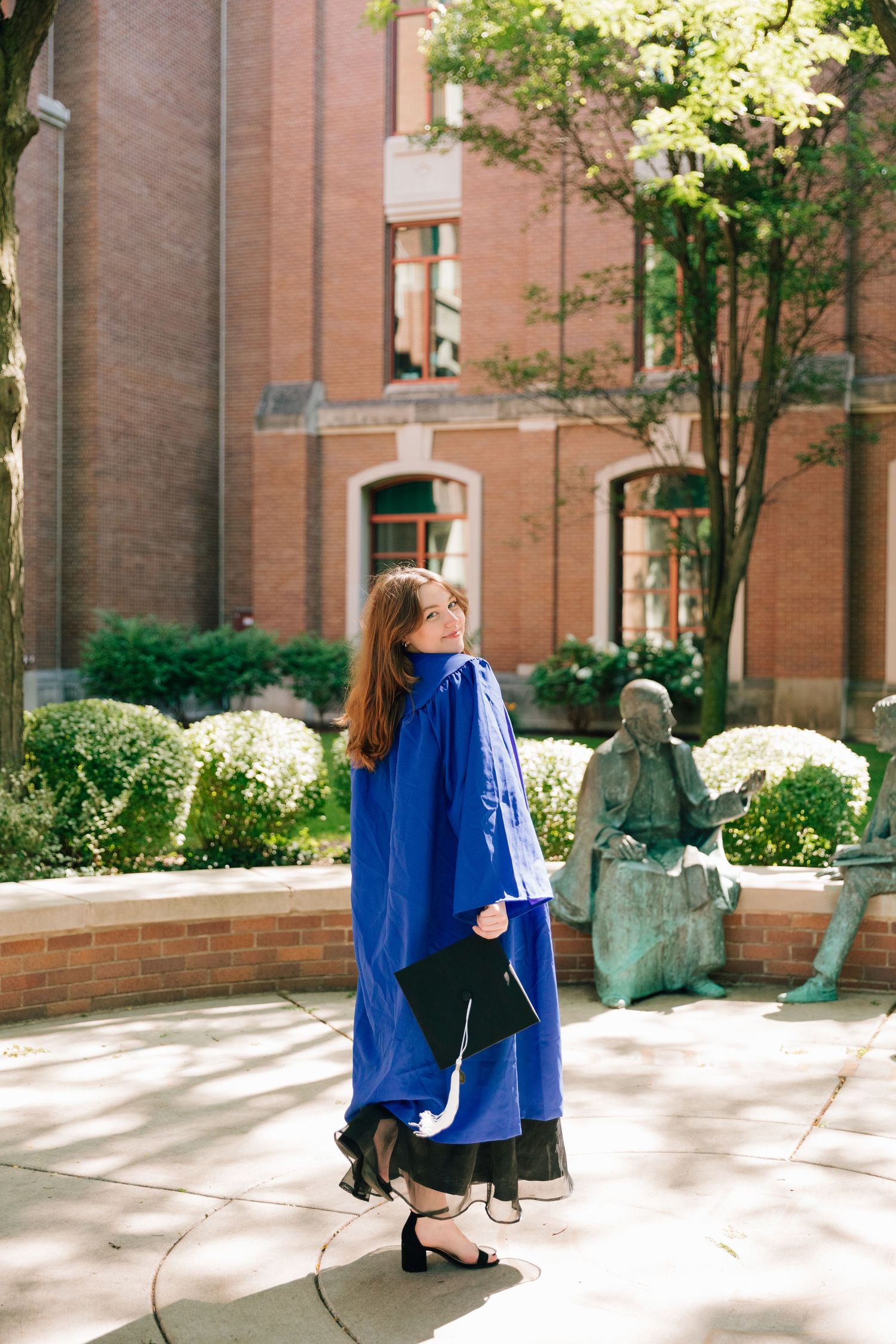 Graduate wearing blue regalia poses next to statue in university courtyard with red brick building.