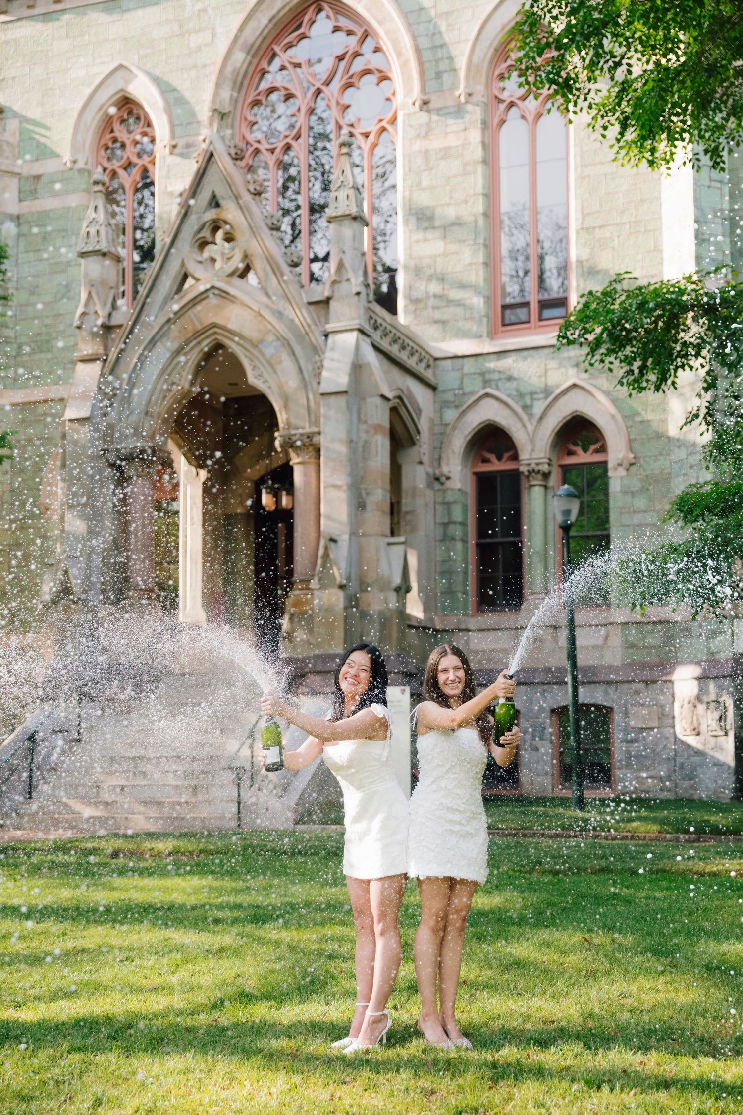 Graduates in white dresses celebrate with champagne spray outside gothic university building.