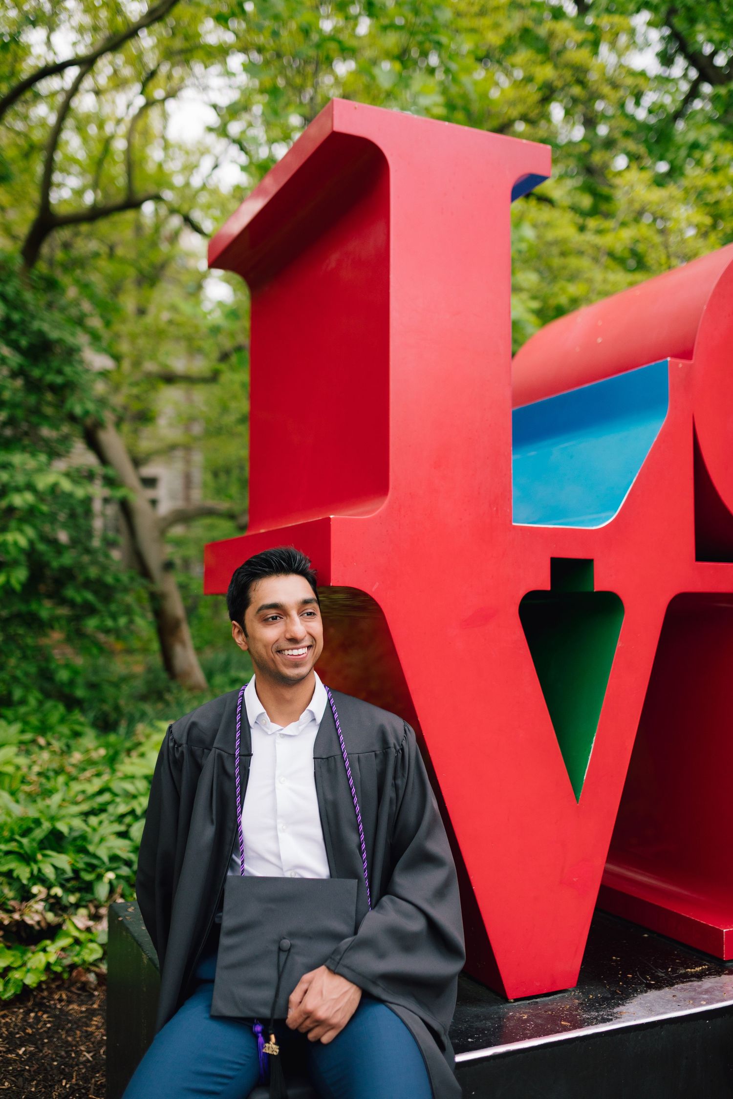 Graduate poses with academic regalia near red LOVE sculpture on campus.