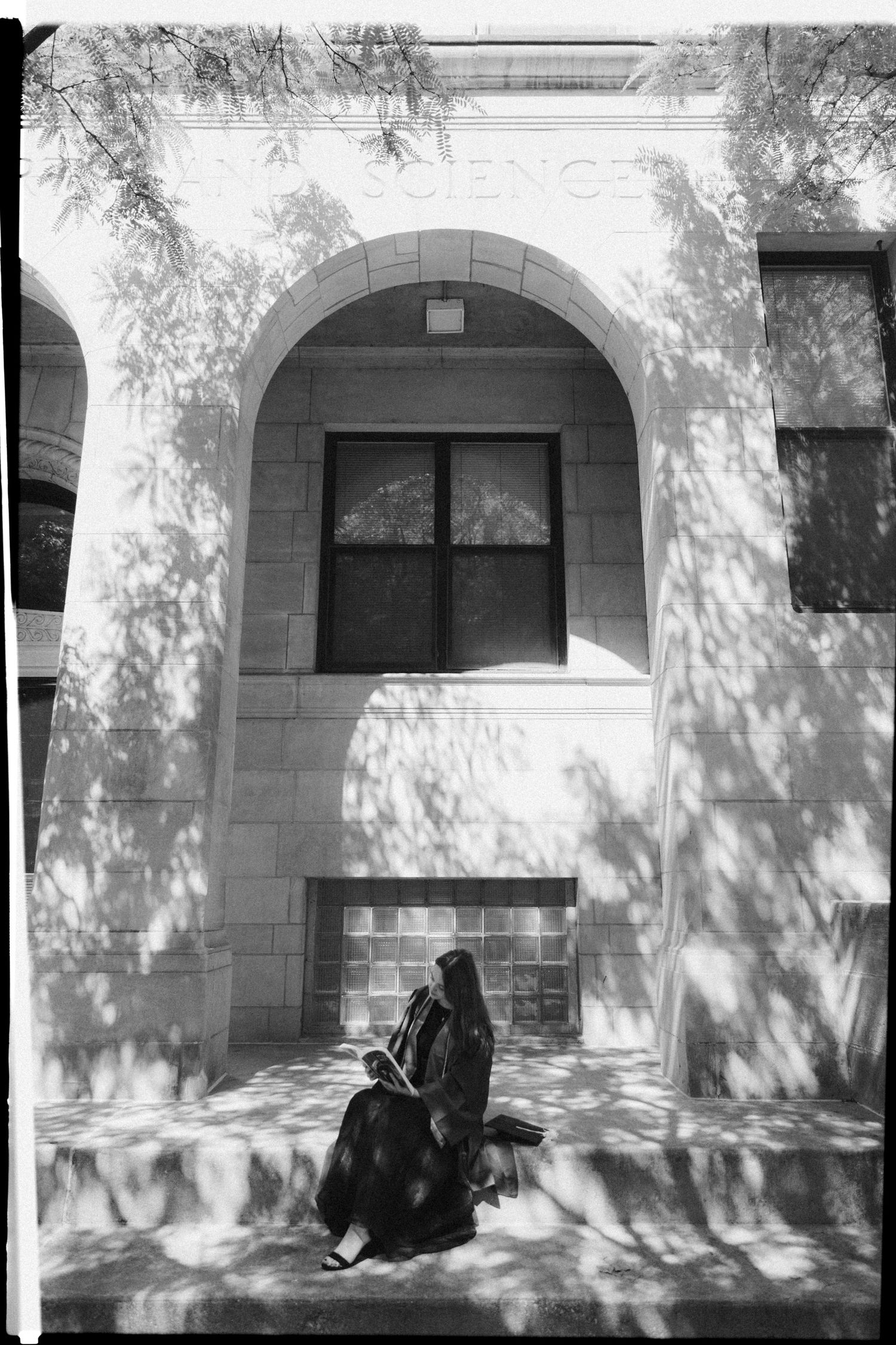 Black and white graduation portrait against stone building with dramatic tree shadows.