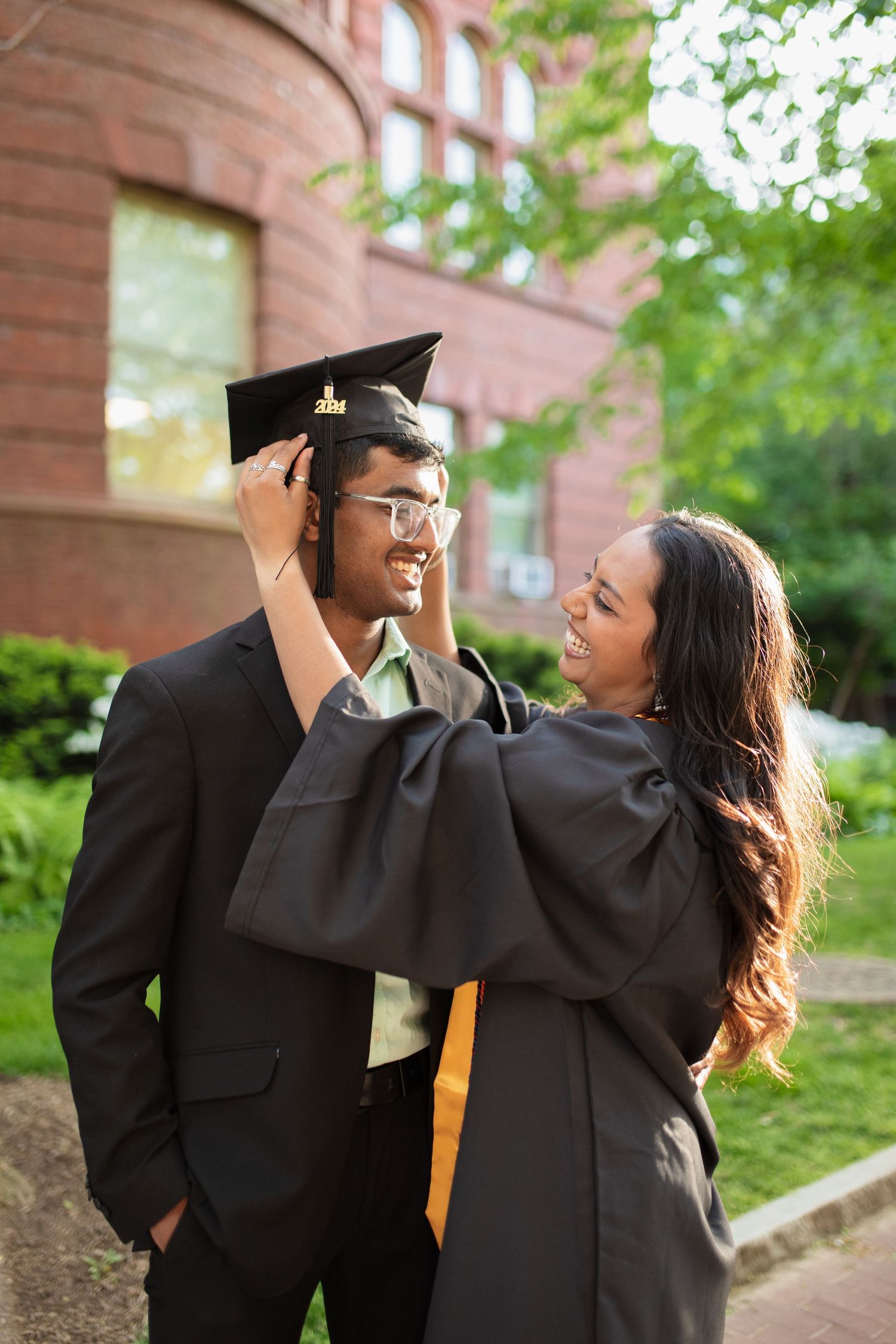 Two graduates share tender moment adjusting graduation cap outside brick building.