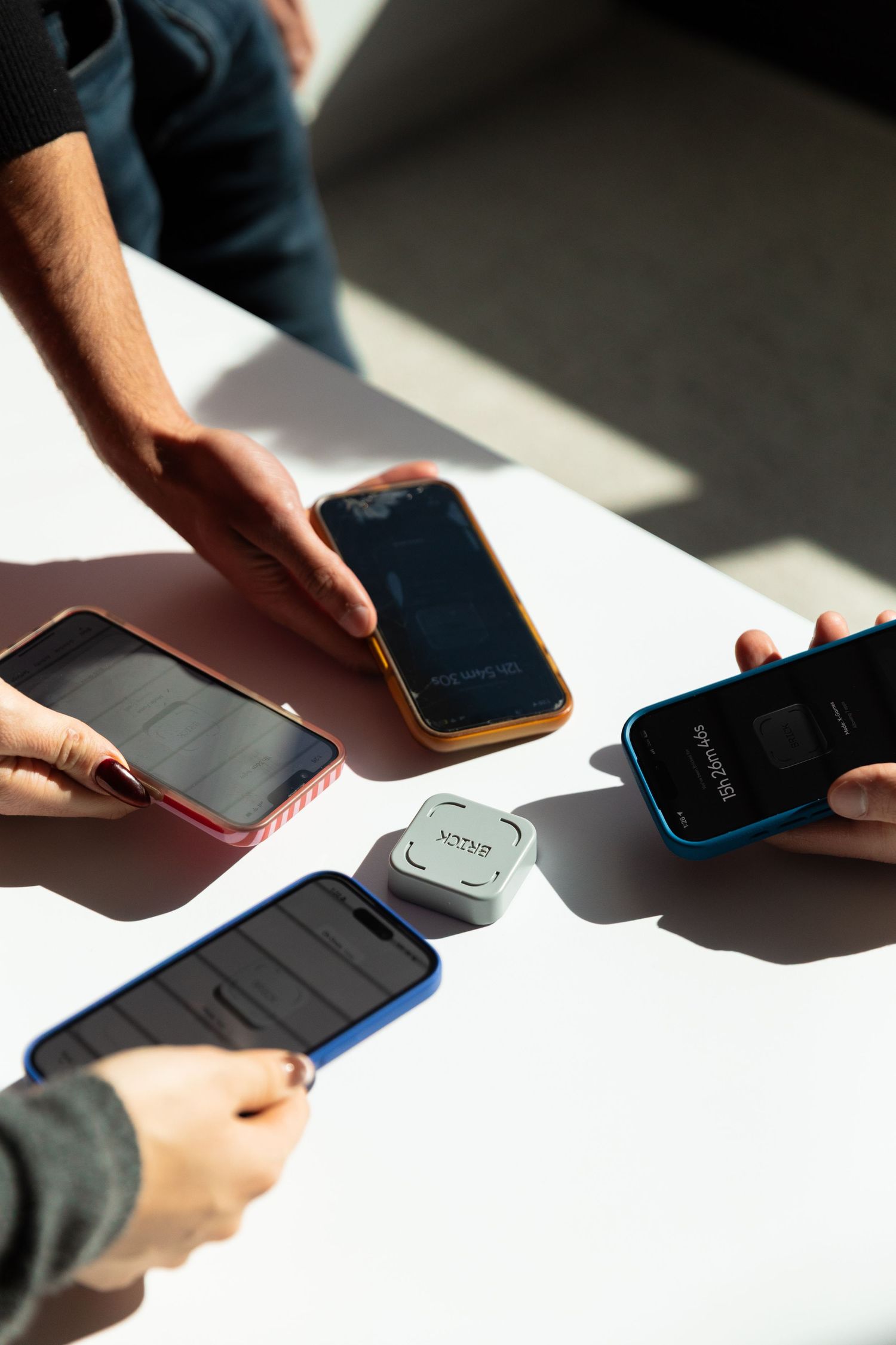 Multiple hands holding smartphones around a small square NFC device on a white table.