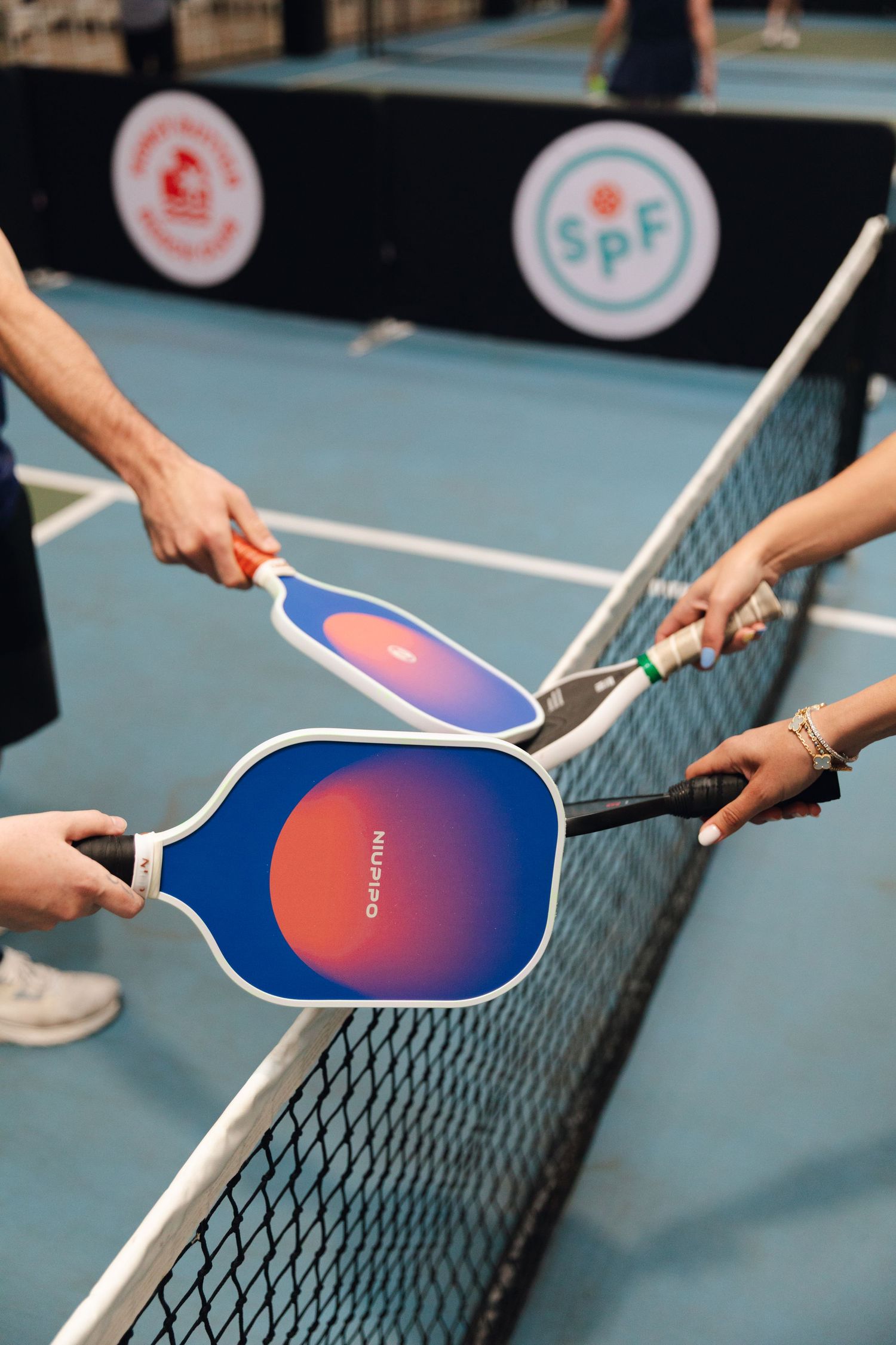 Players tap pickleball paddles over the net in a post-game handshake on a blue indoor court.