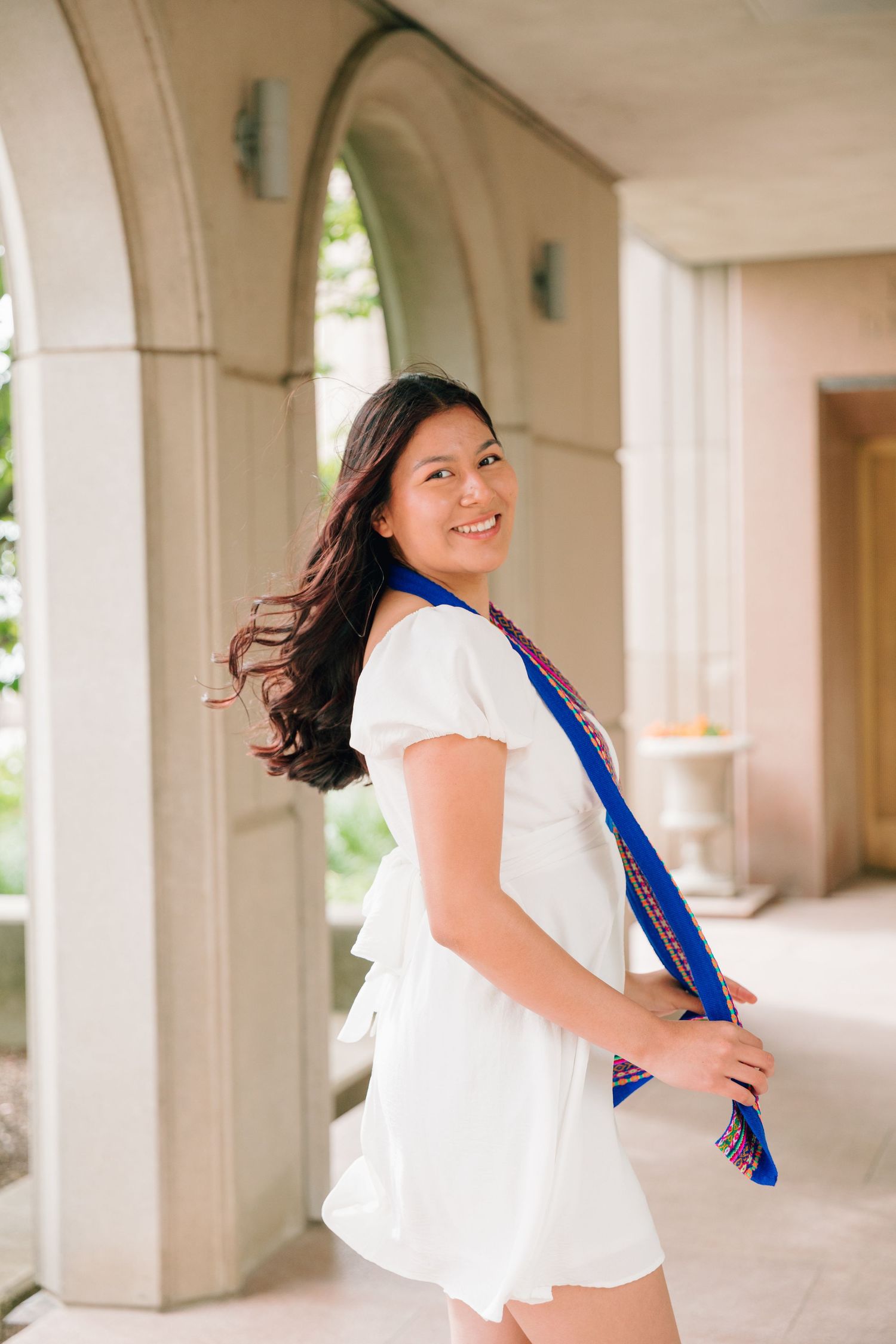 Graduate in white dress with blue sash poses in columned hallway.