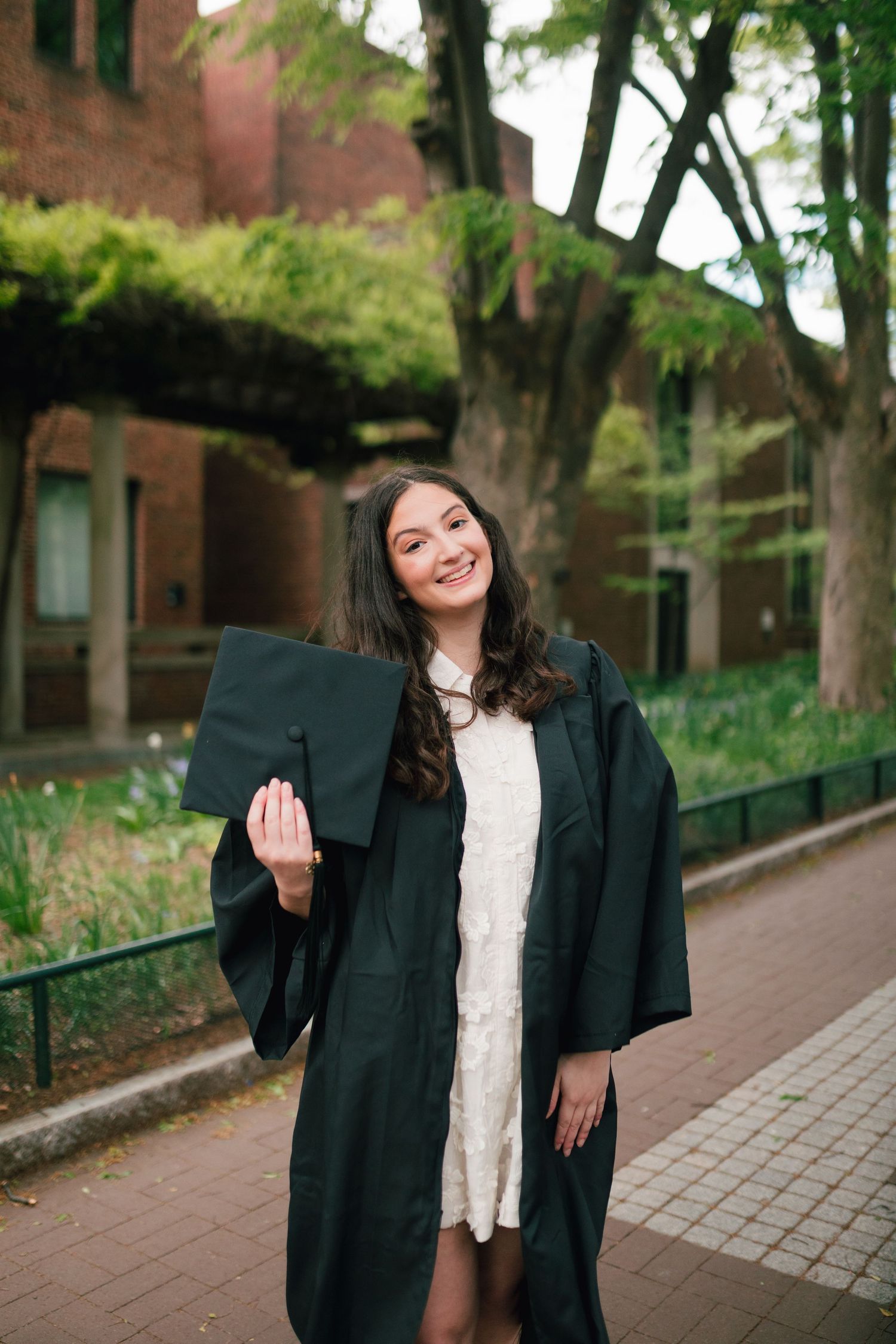 Graduate in academic regalia poses on brick pathway surrounded by greenery.