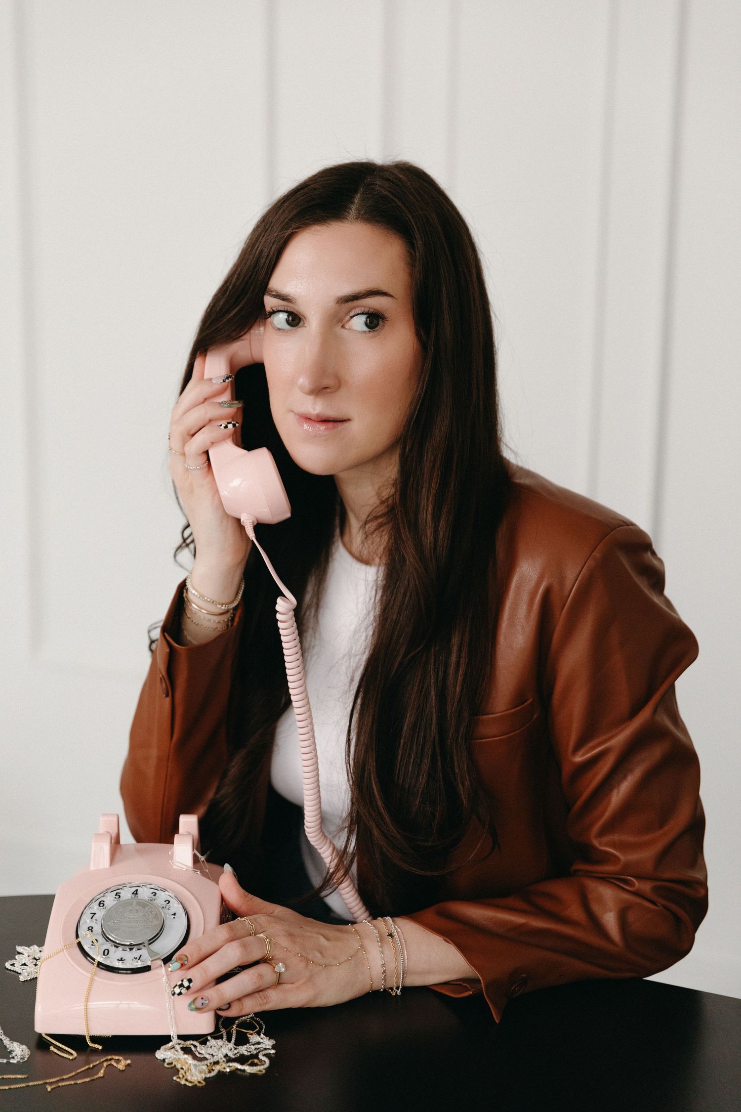 Woman in brown leather jacket talking on a vintage pink rotary phone at a dark table with jewelry.