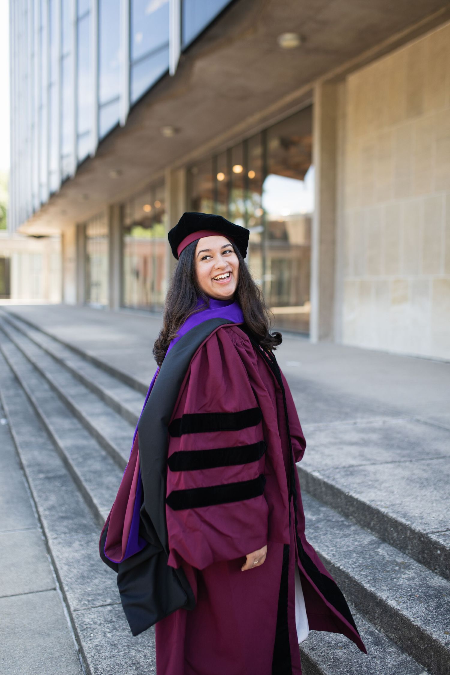 Graduate in purple doctoral regalia smiles while posing on concrete steps.