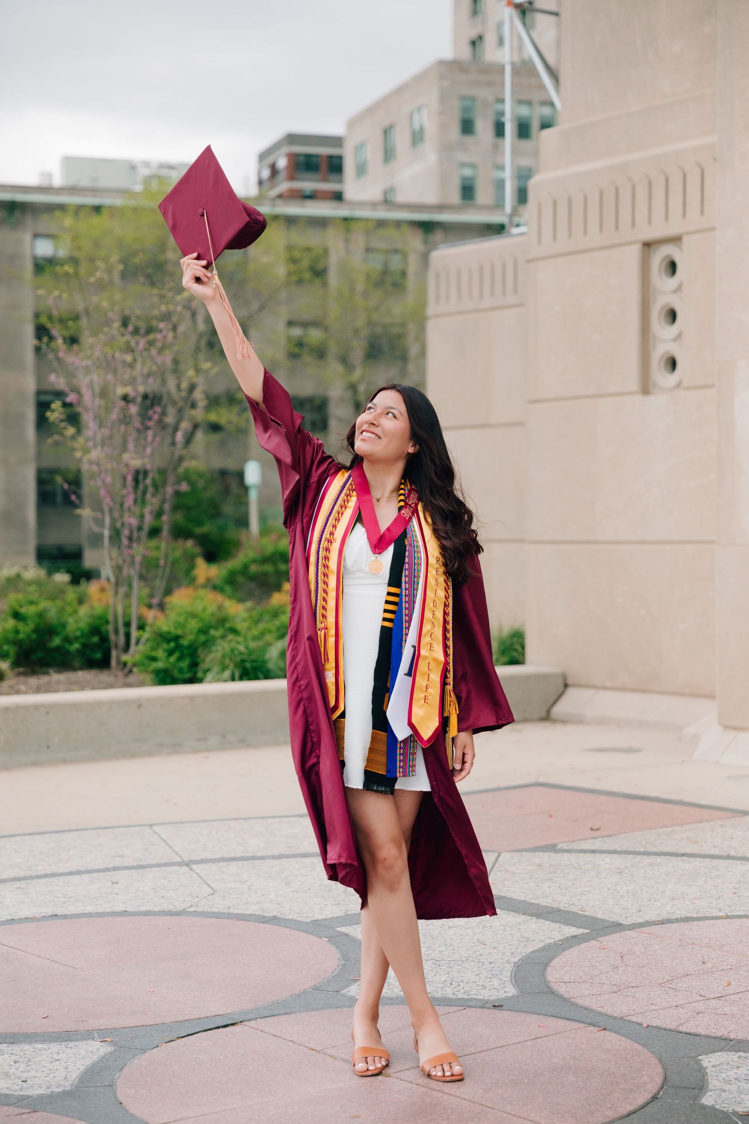 Graduate in maroon graduation gown holds cap high while standing on decorative circular pavement.