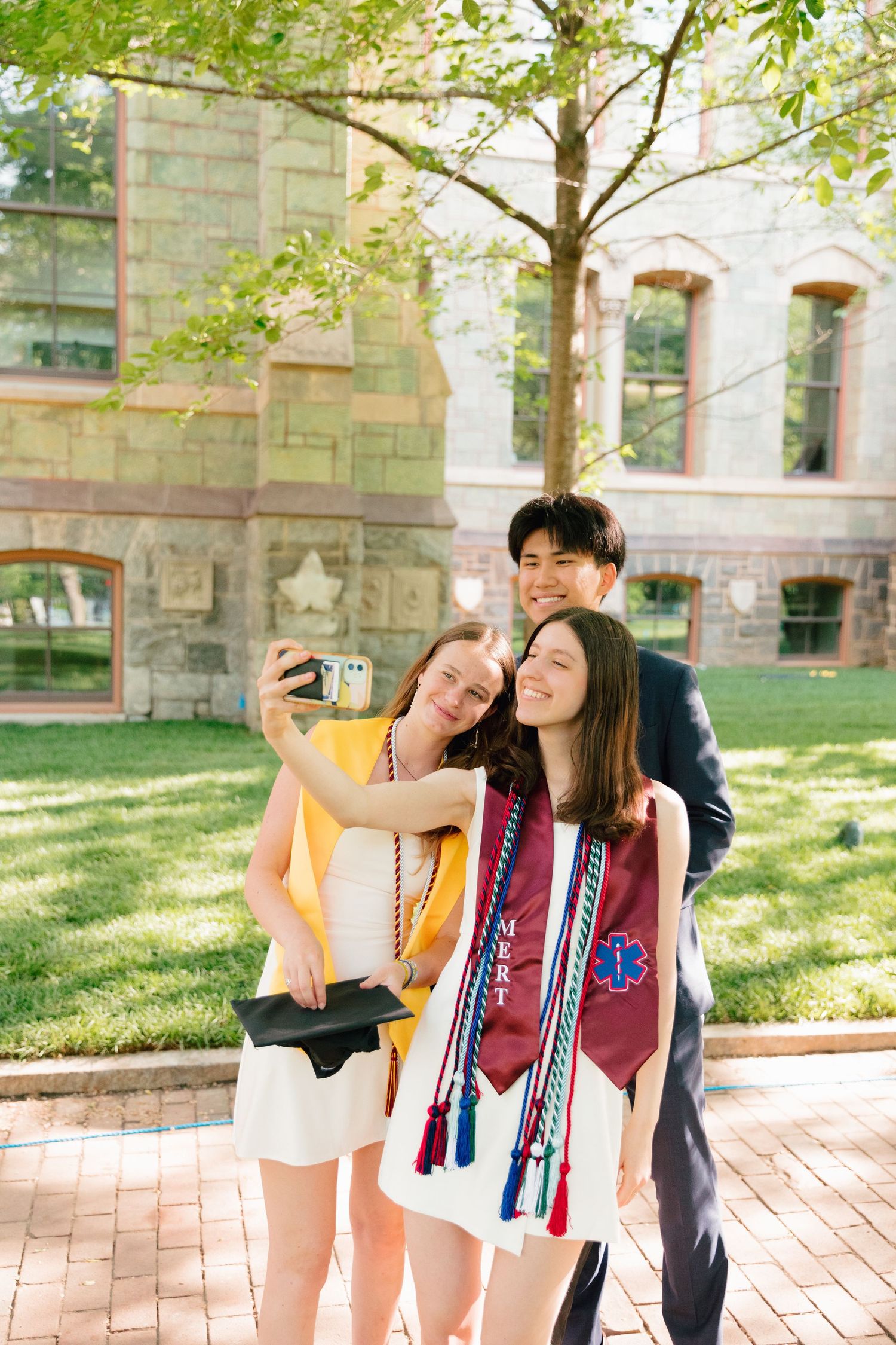 Three graduates take selfie together on sunny university campus.