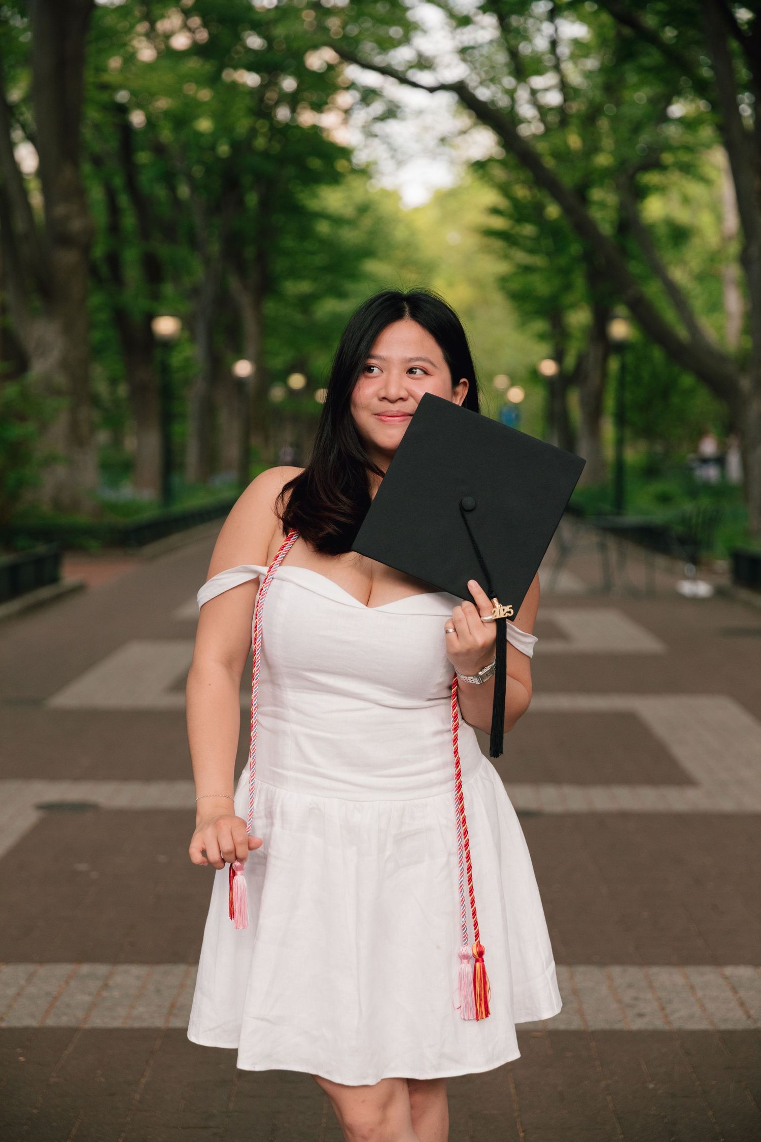 Graduate celebrates on campus pathway wearing white dress and graduation cap.