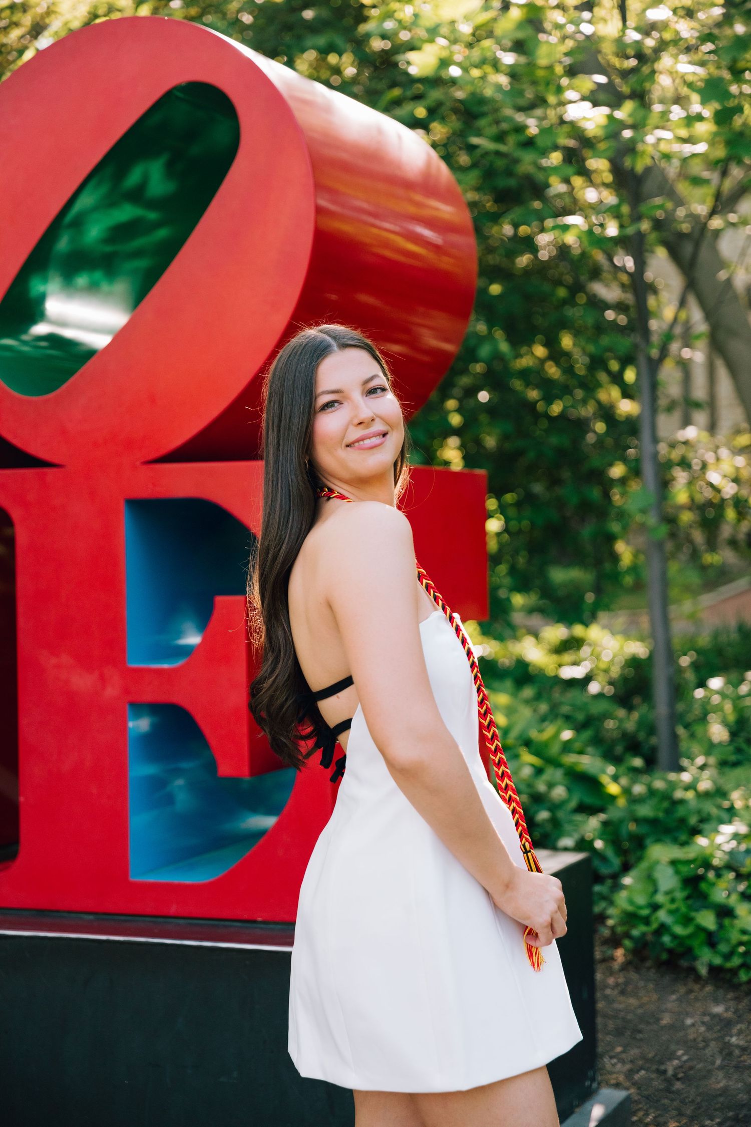 Graduate in white dress poses joyfully by red LOVE sculpture outdoors.