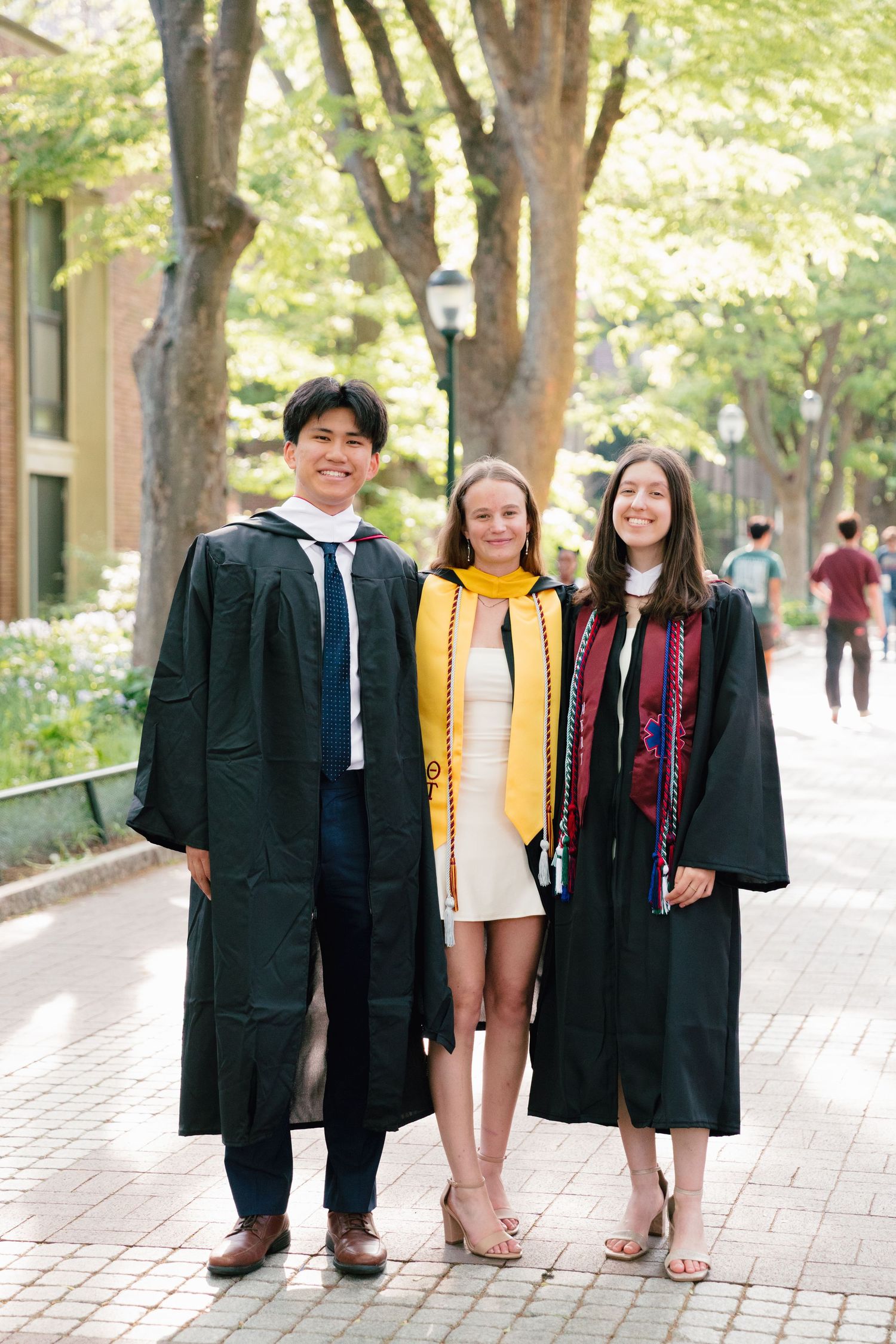 Three graduates in academic regalia pose on tree-lined campus pathway.