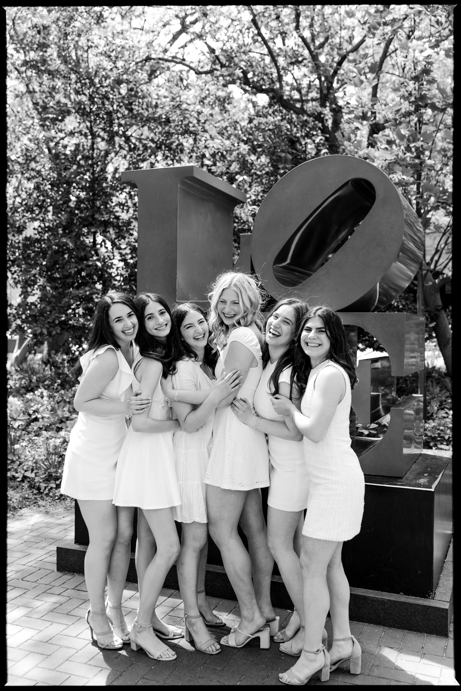 A group poses in white dresses next to a large black sculpture outdoors.