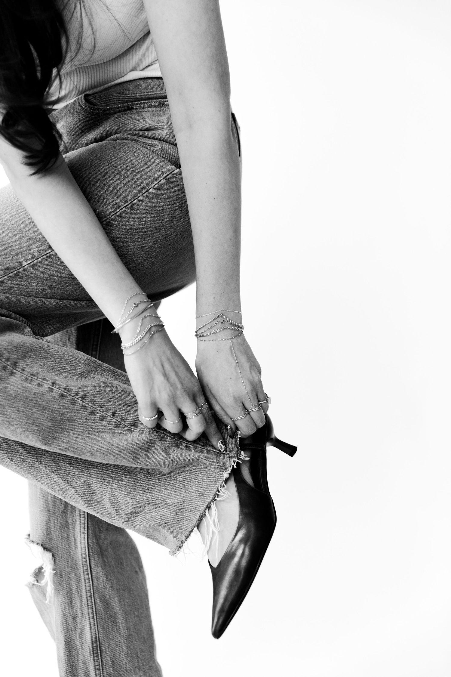 Woman in distressed jeans adjusting a black pointed-toe kitten heel shoe, black and white photo.