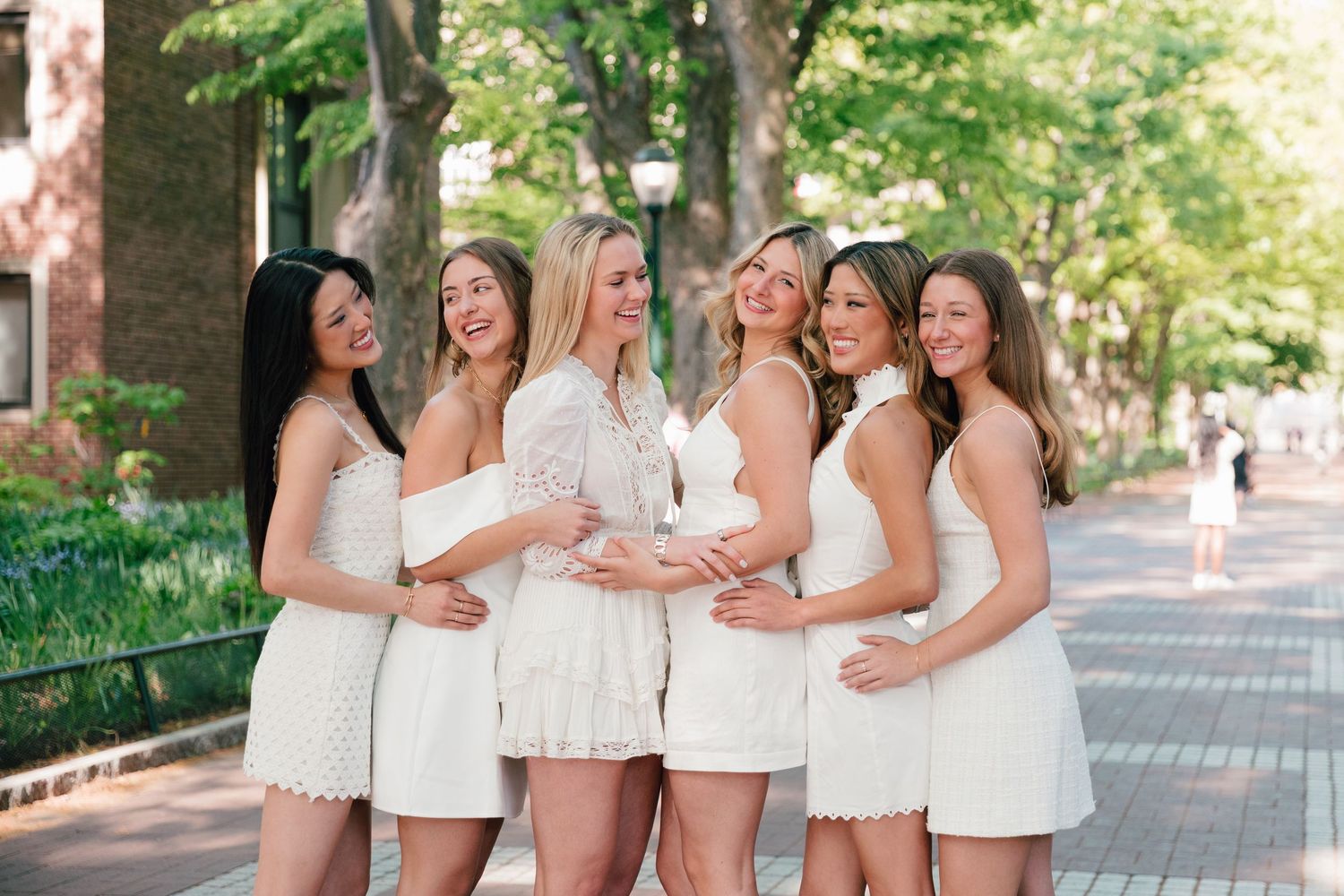 Group of graduates in white dresses celebrate together on a scenic campus path.