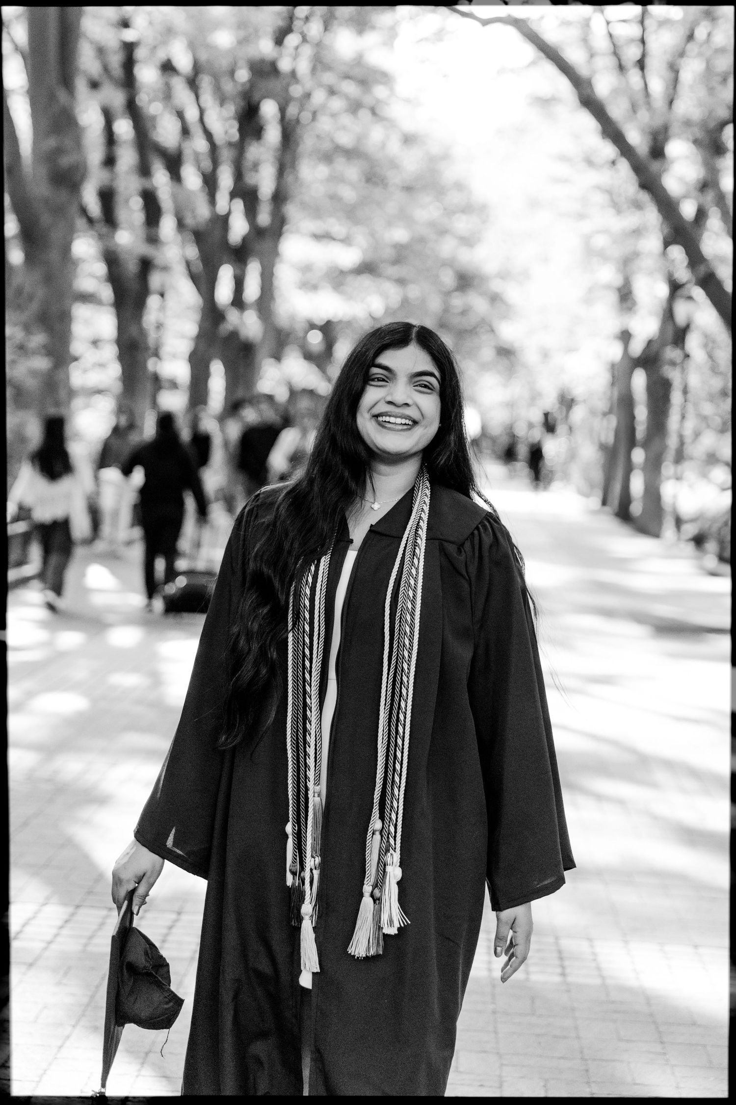 Graduate in academic regalia smiling on tree-lined campus path in black and white photo.
