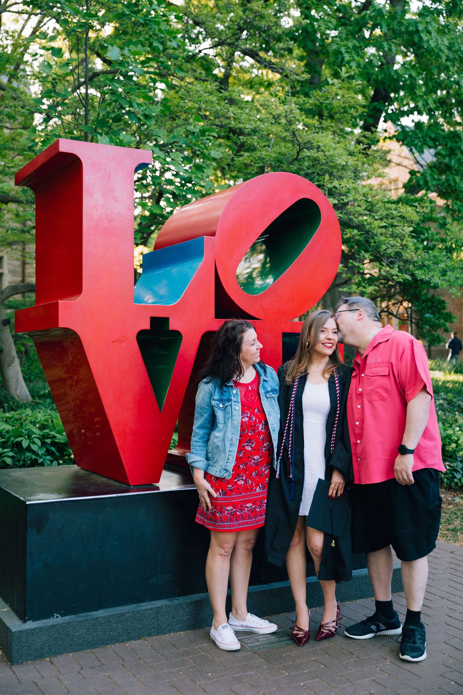 Three people pose playfully by red LOVE sculpture in park setting.