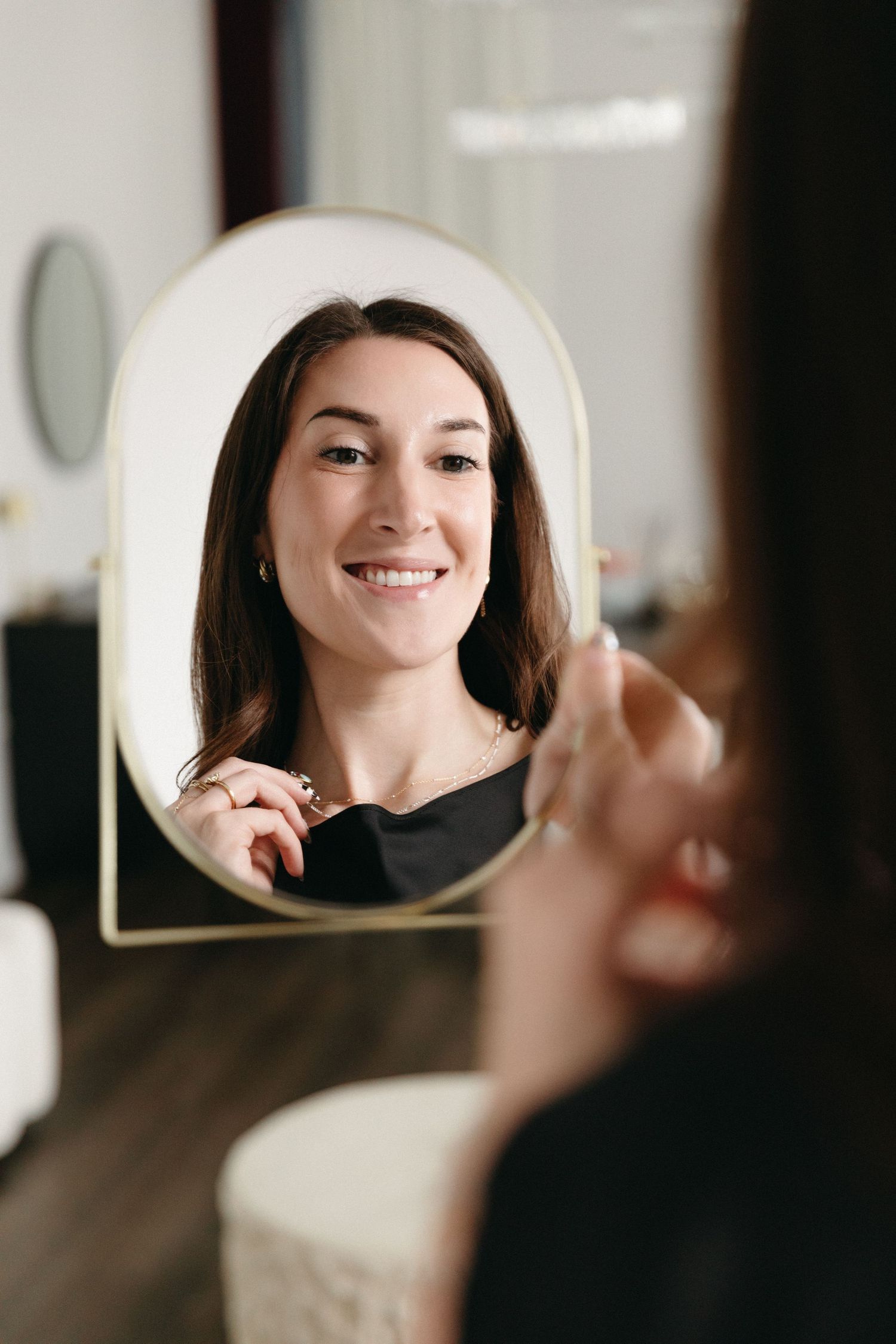Smiling brunette woman admiring her reflection in a gold oval mirror while touching her face indoors.
