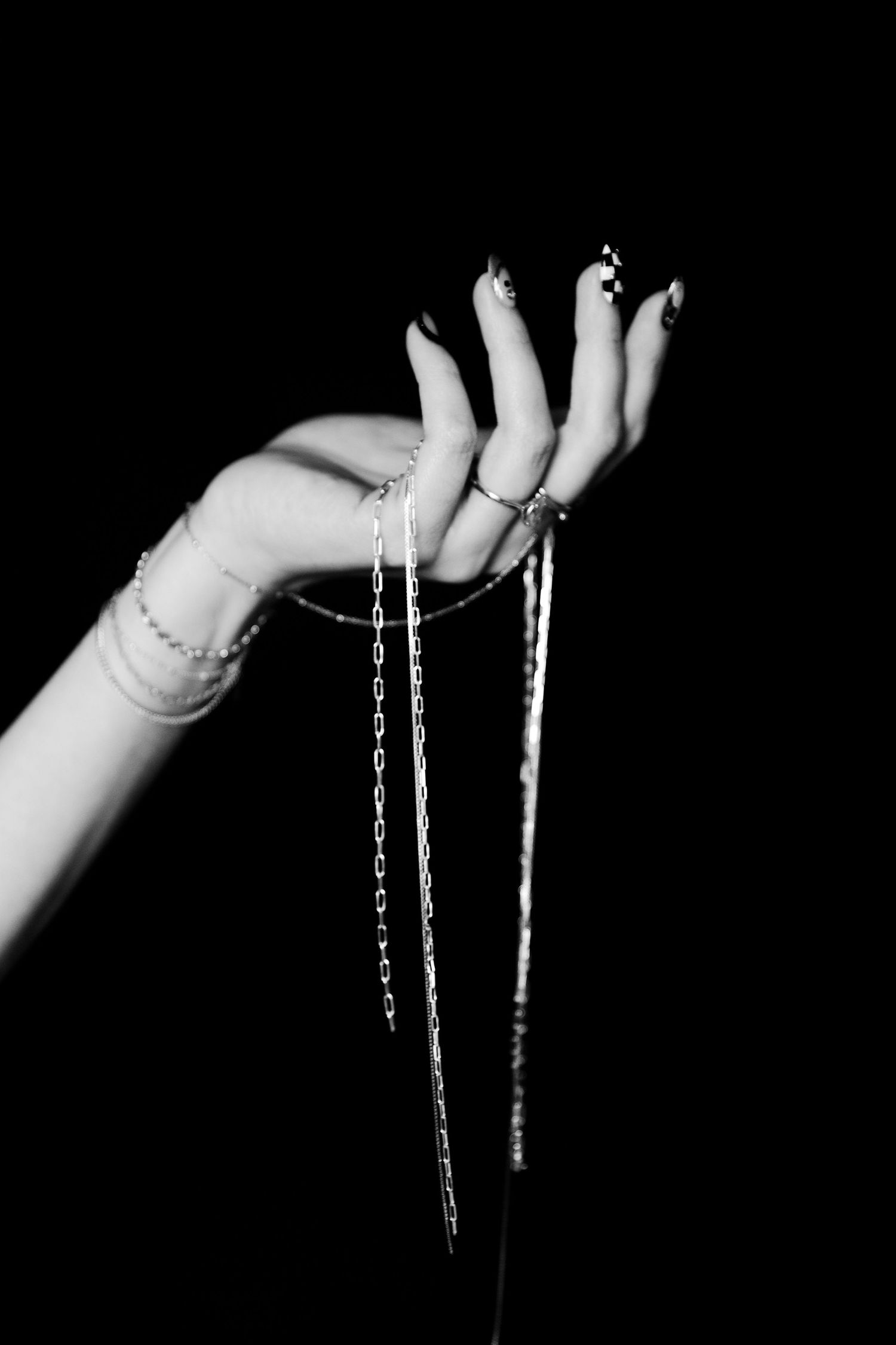 Black and white photo of a hand with dark nails and rings holding delicate silver chains against a black background.