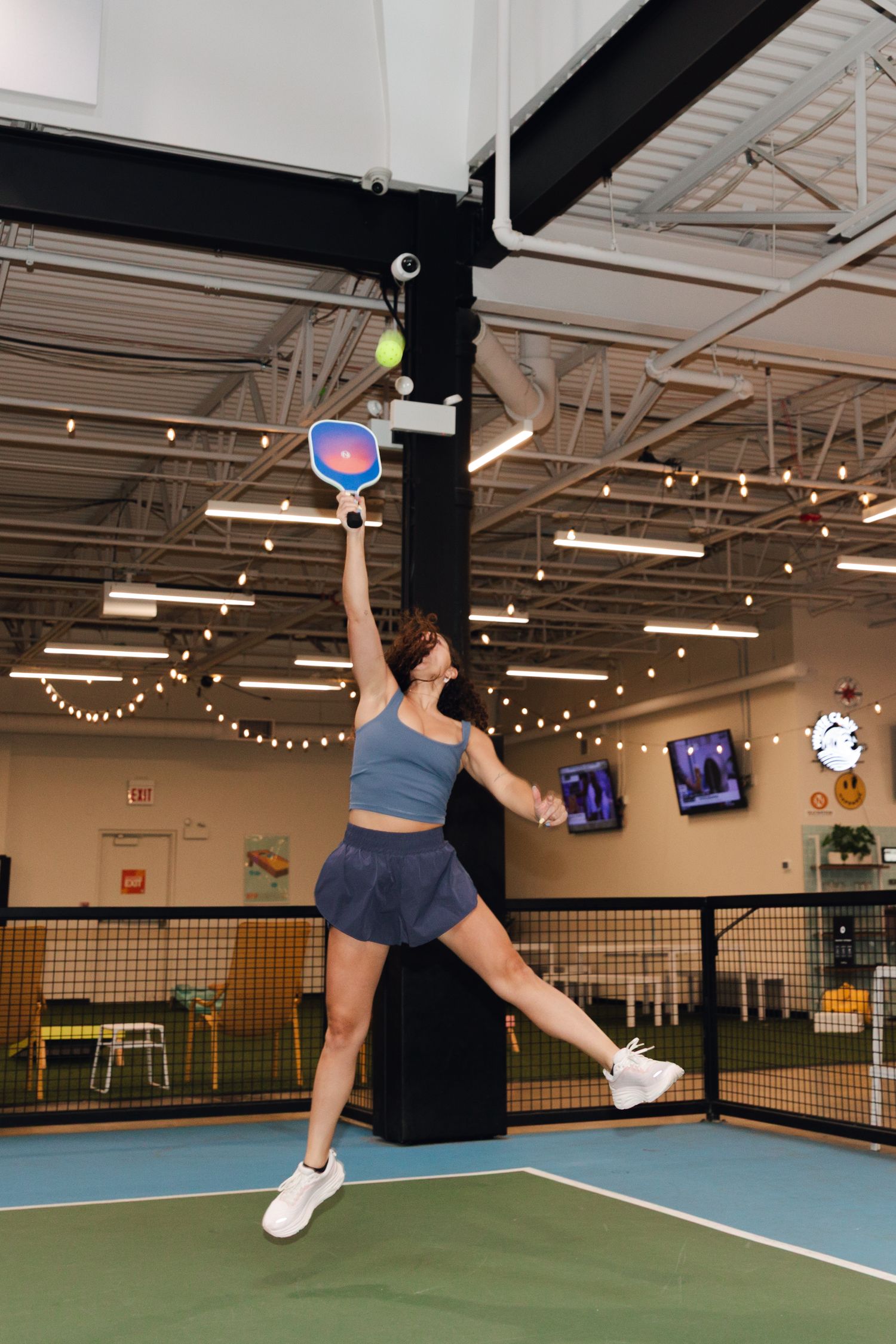 Woman jumping mid-air on a pickleball court, reaching high with a blue paddle to hit a ball indoors.