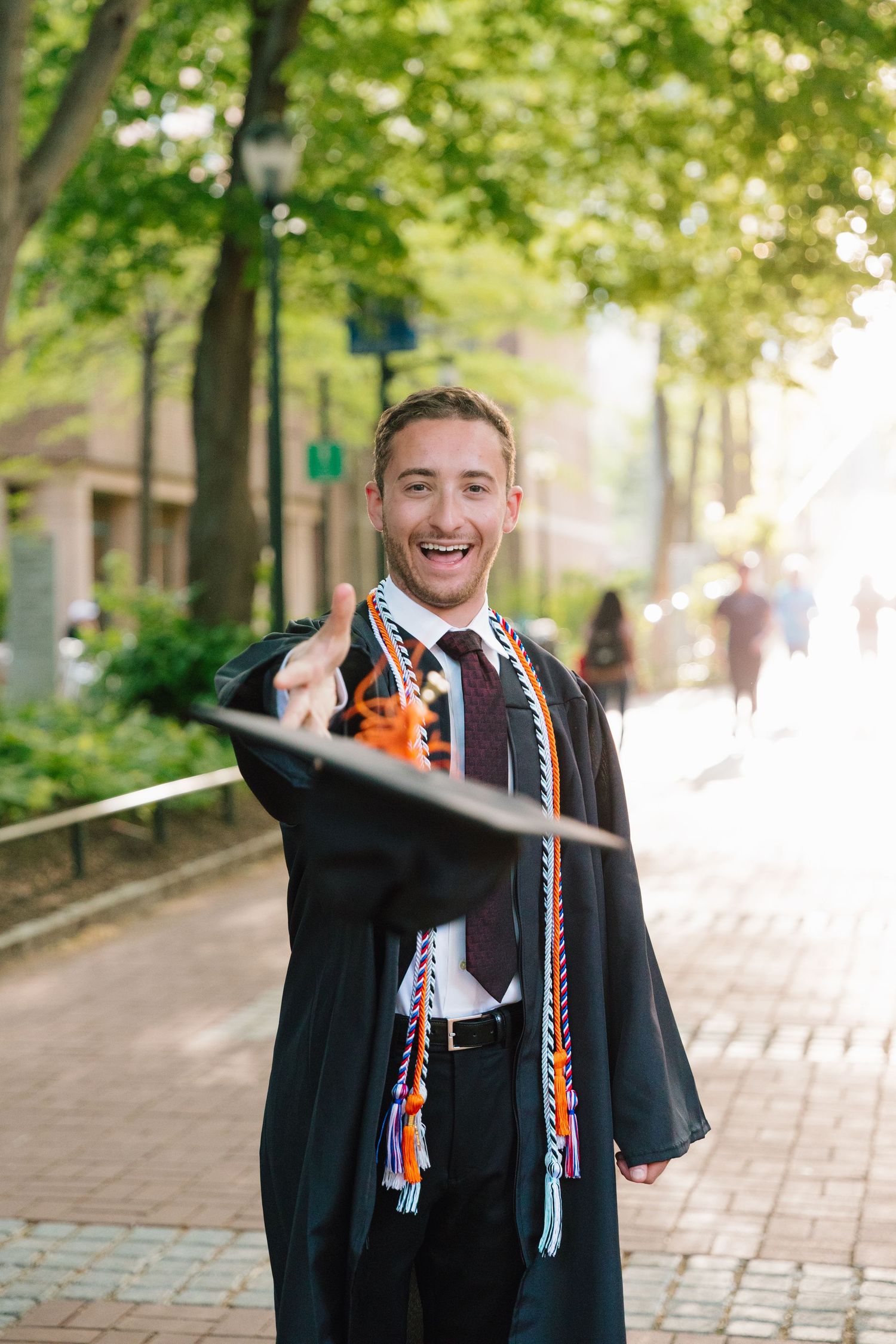 Graduate in black academic regalia with colorful cords poses on a tree-lined campus pathway.