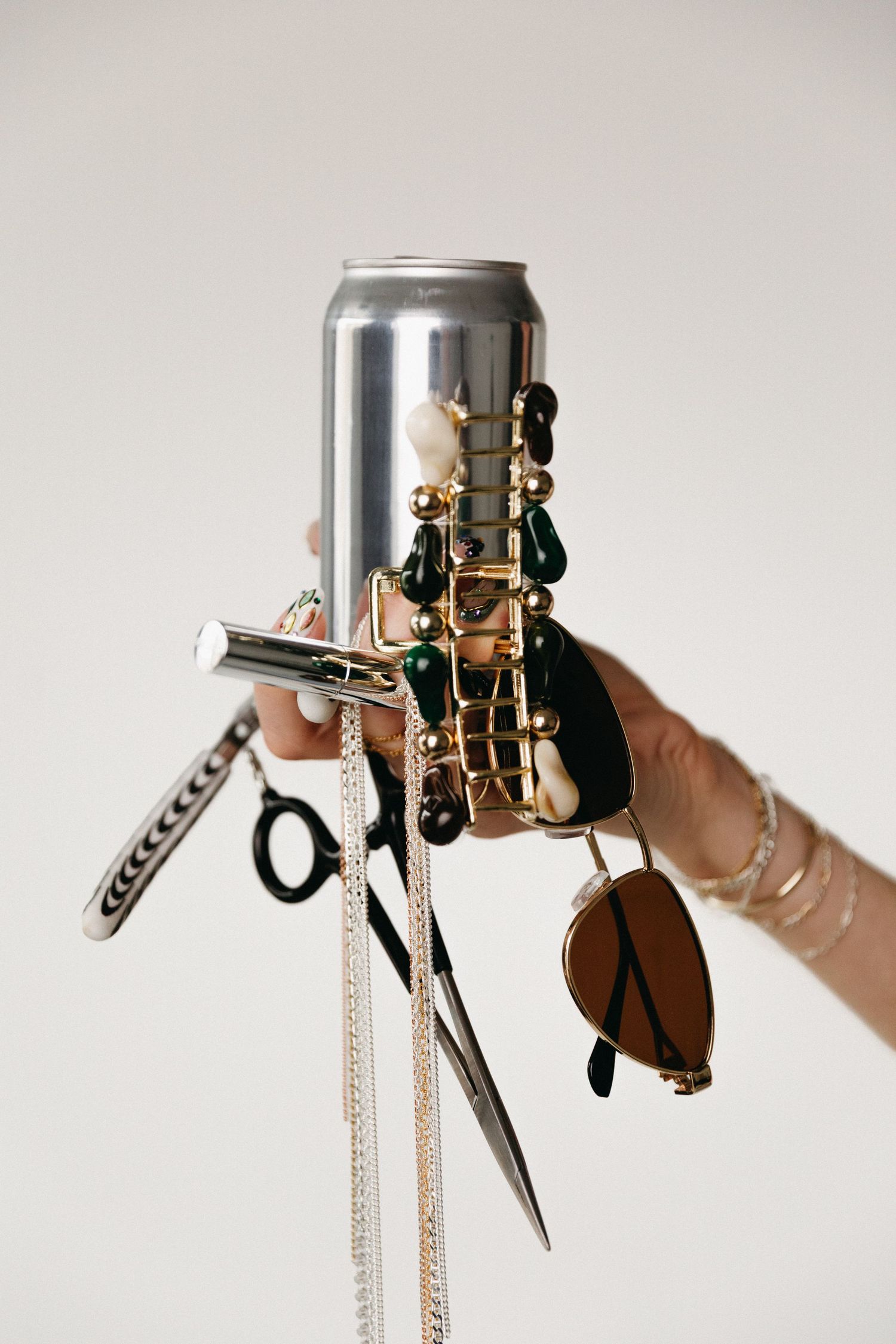 A hand adorned with gold rings and bracelets holds a silver can alongside scissors, sunglasses, and jewelry accessories.