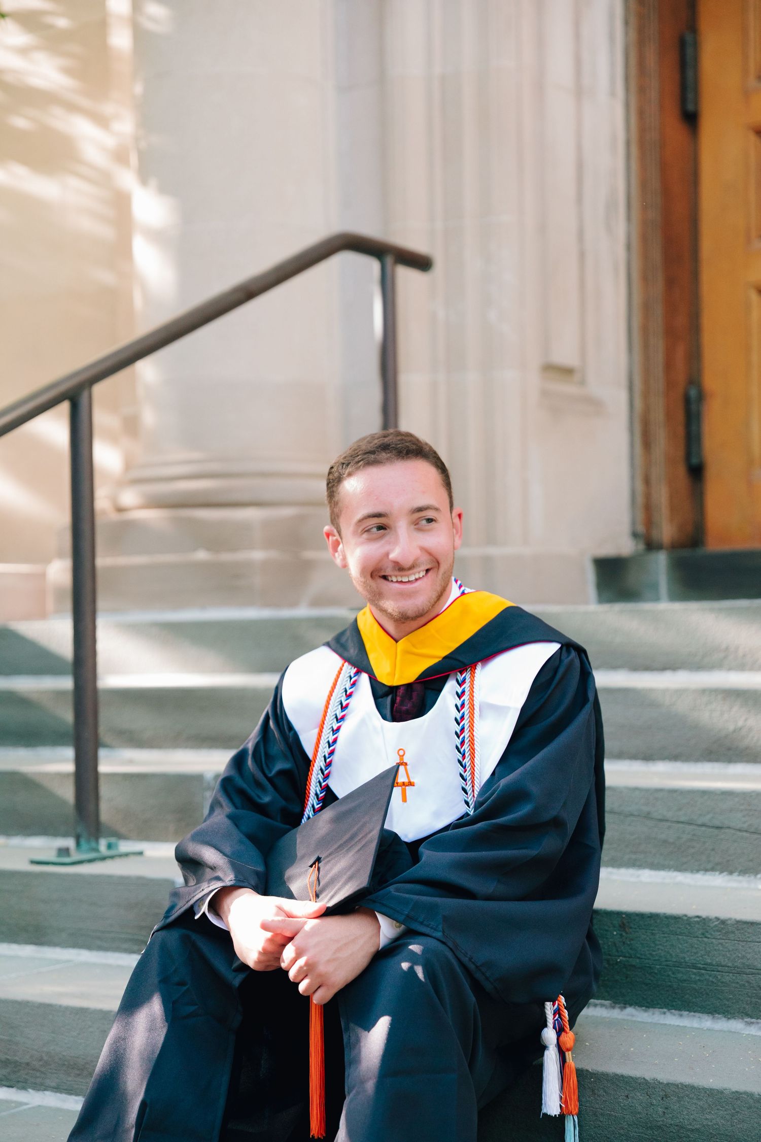 Graduate in academic regalia with yellow hood sitting on stone steps.