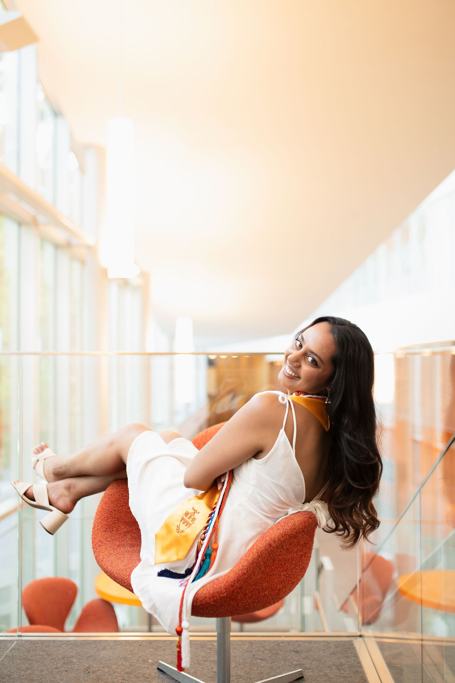 Graduate in white dress reclines in orange modern chair for stylish portrait.