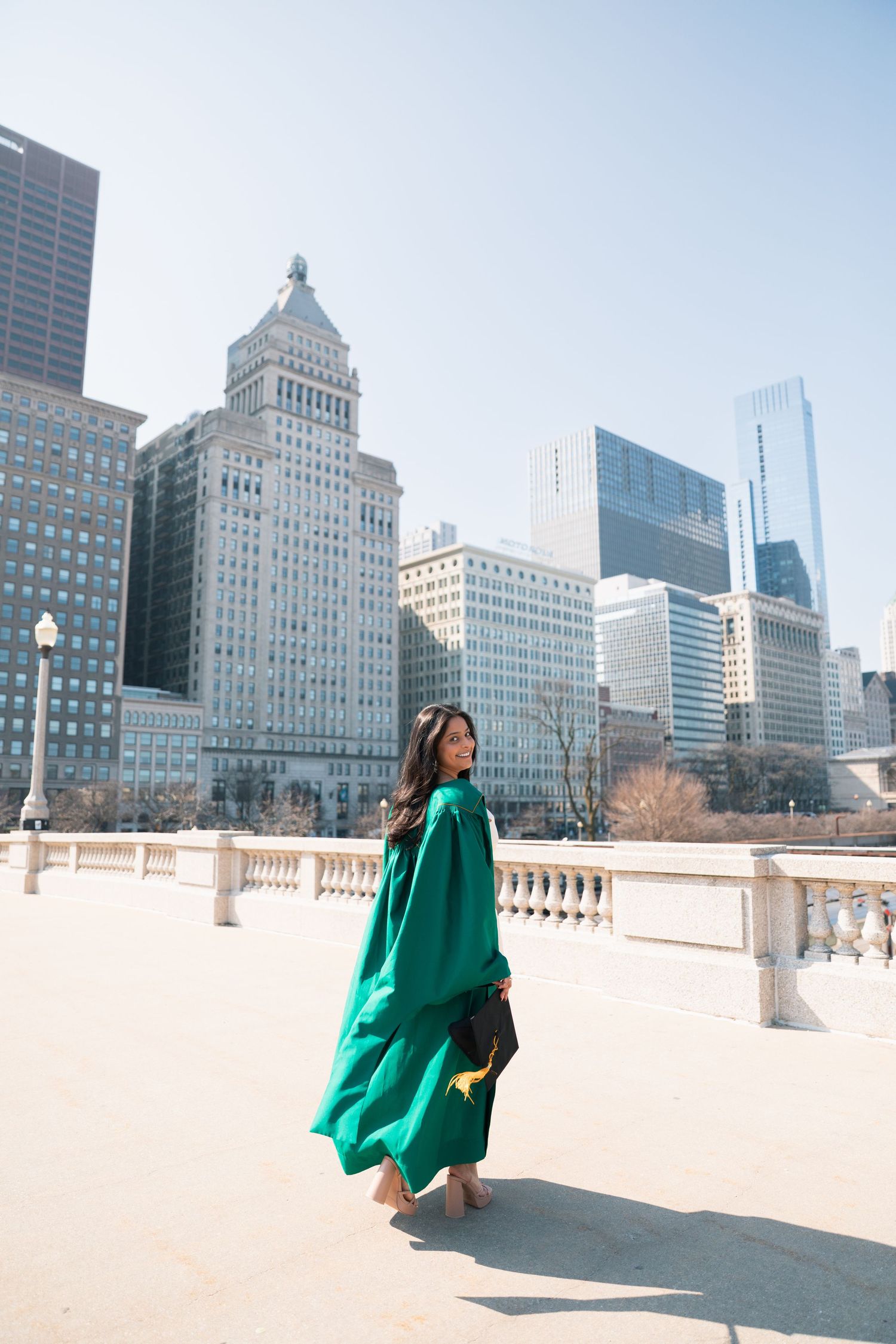 Green-robed graduate walks along Chicago's Grant Park with skyline backdrop.