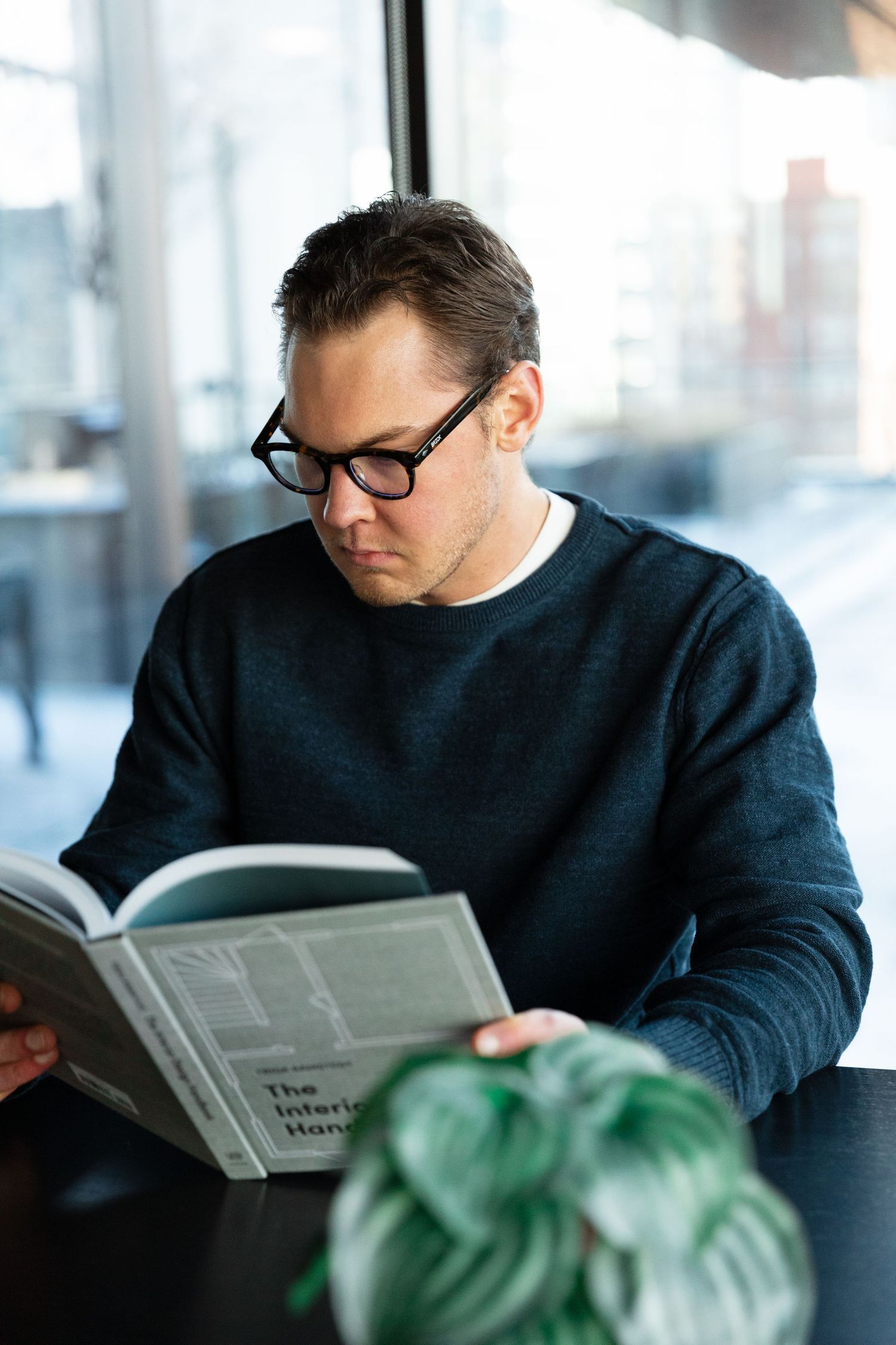 Man with black glasses reading The Interior Handbook book while sitting at a table near a window.